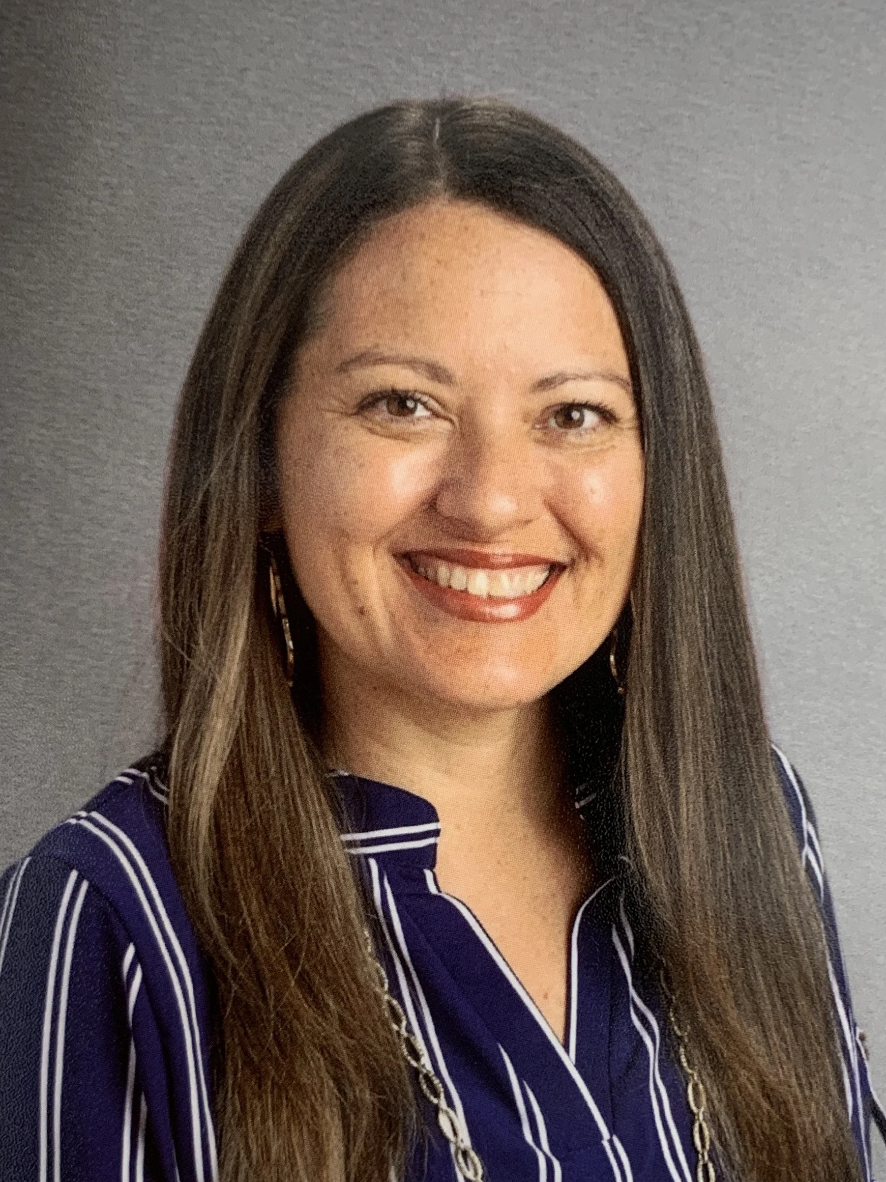 A woman with long brown hair smiling, wearing a navy blue and white striped shirt, earrings, and a gold chain necklace.