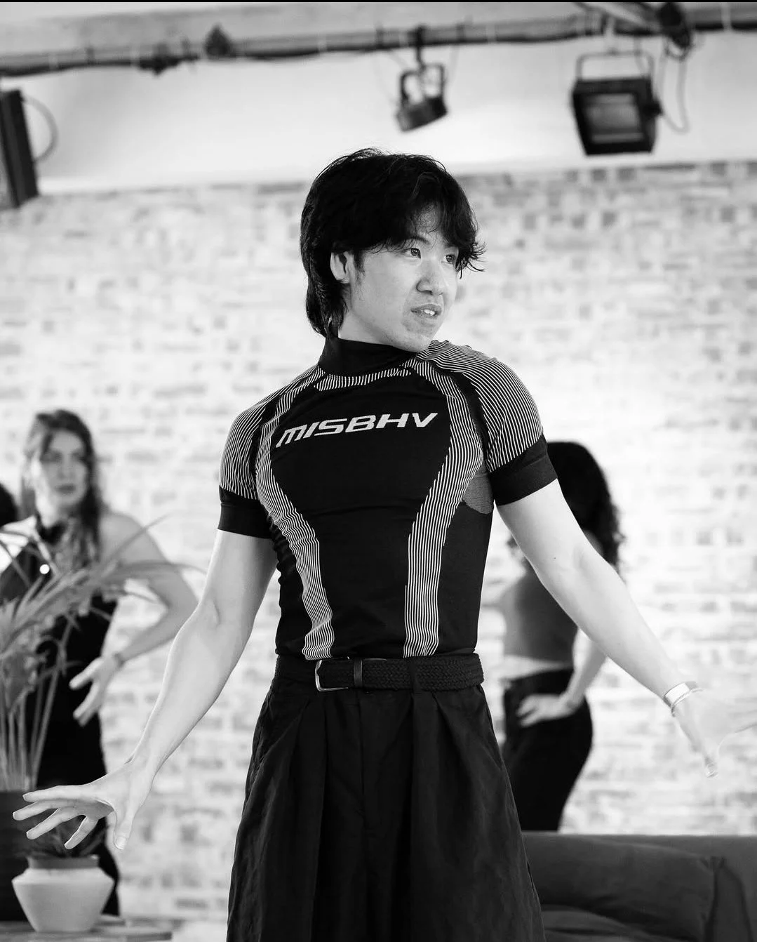 A man wearing a sports shirt with the text 'MISBHV' on it, standing indoors with a brick wall background. Two women are visible in the background.