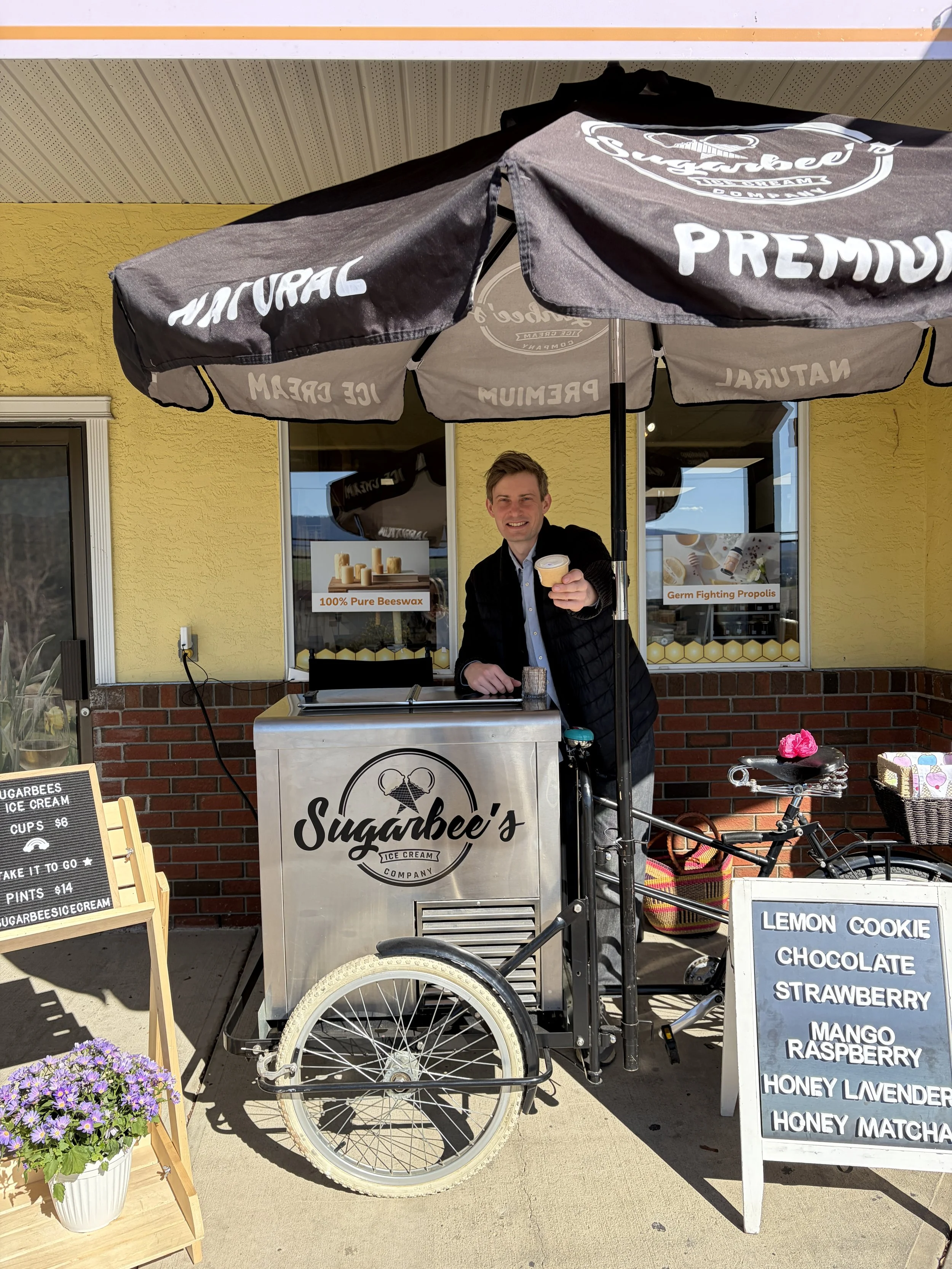 A man standing behind a sugarbee's ice cream cart under a black umbrella saying 'Natural' and 'Premium' in front of a yellow building with posters about beeswax and germ fighting propolis. The cart has a bicycle wheel and a small chalkboard menu listing flavors including lemon cookie, chocolate, strawberry, mango, raspberry, honey lavender, and honey matcha. There's a potted purple flower on the sidewalk.