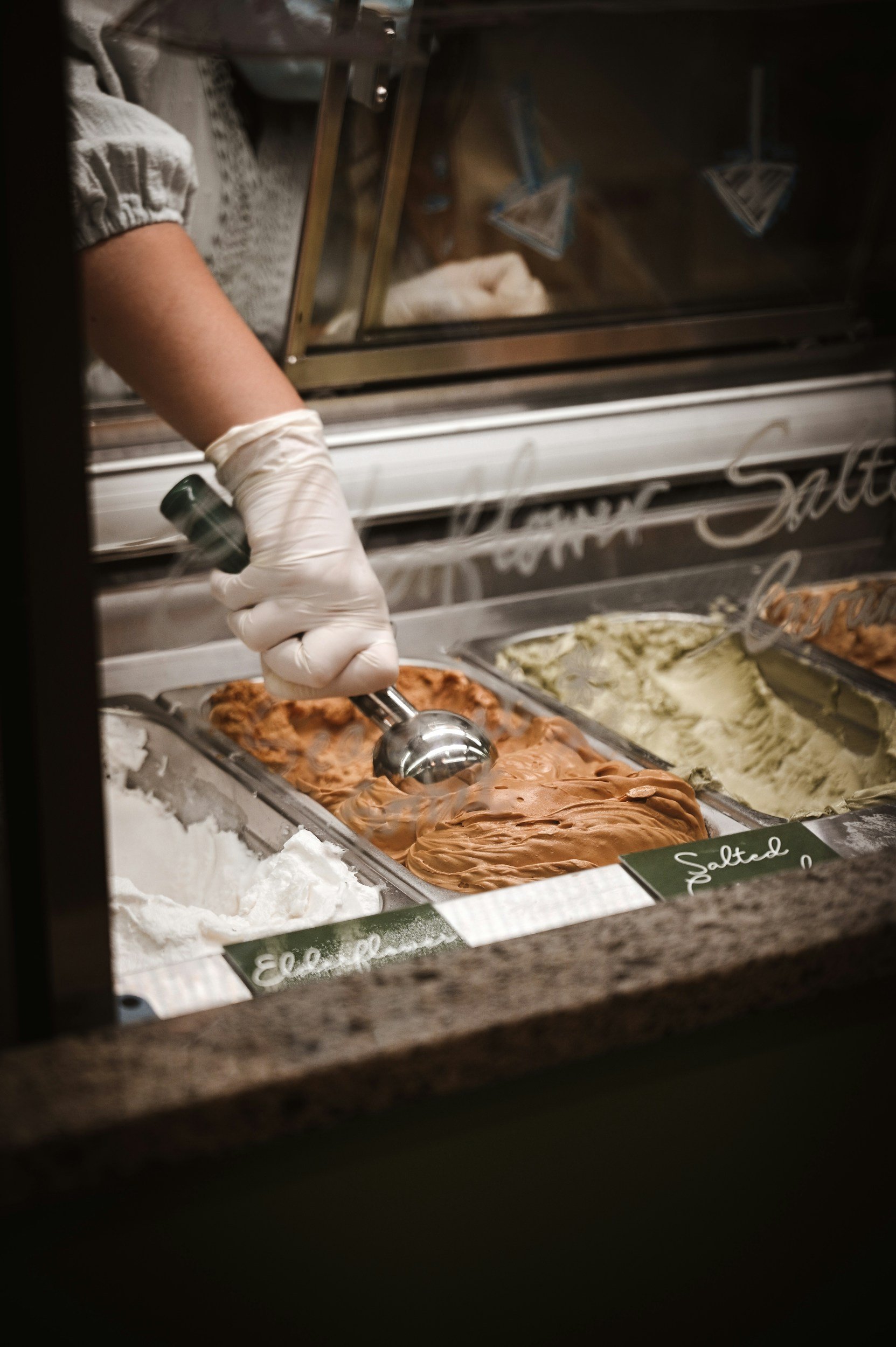 Ice cream shop with various flavors, including salted caramel, in stainless steel containers, with a hand in a glove scooping ice cream.