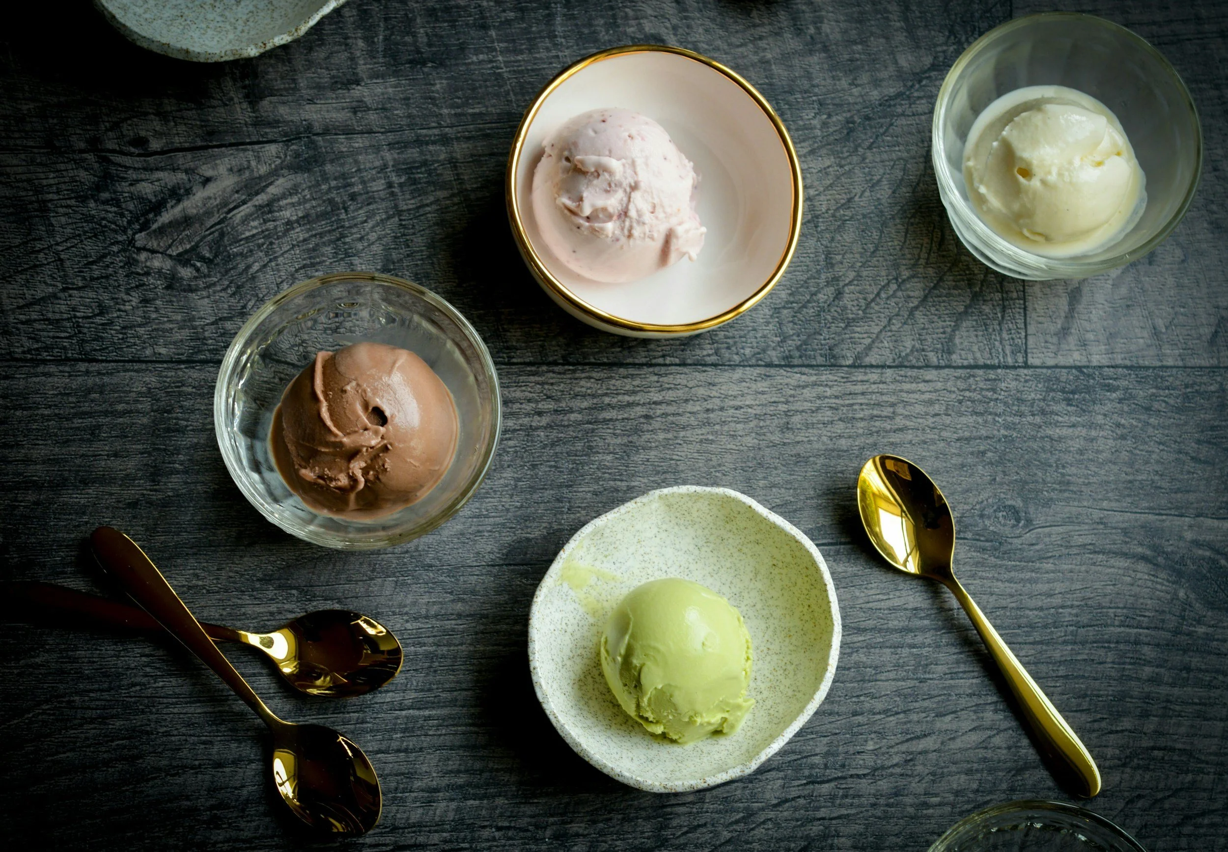 Four bowls of different flavors of ice cream on a dark wooden table, with golden spoons placed next to each bowl.