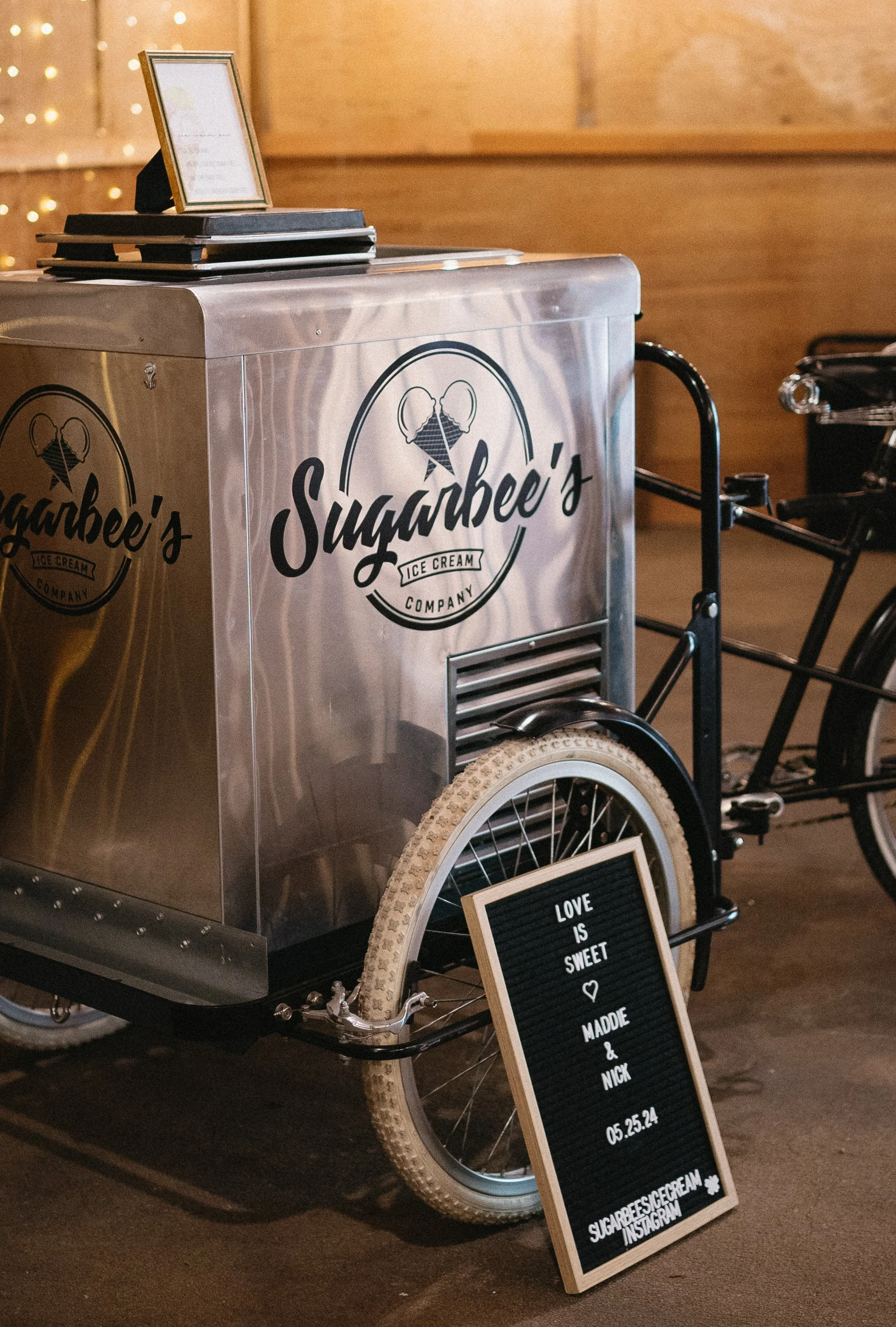 A mobile ice cream cart with the logo 'Sugarbee's Ice Cream Company' and a small chalkboard sign that reads 'Love is sweet, made & nick 05.25.24'. The cart has a large wheel and is parked on a floor with wooden background.