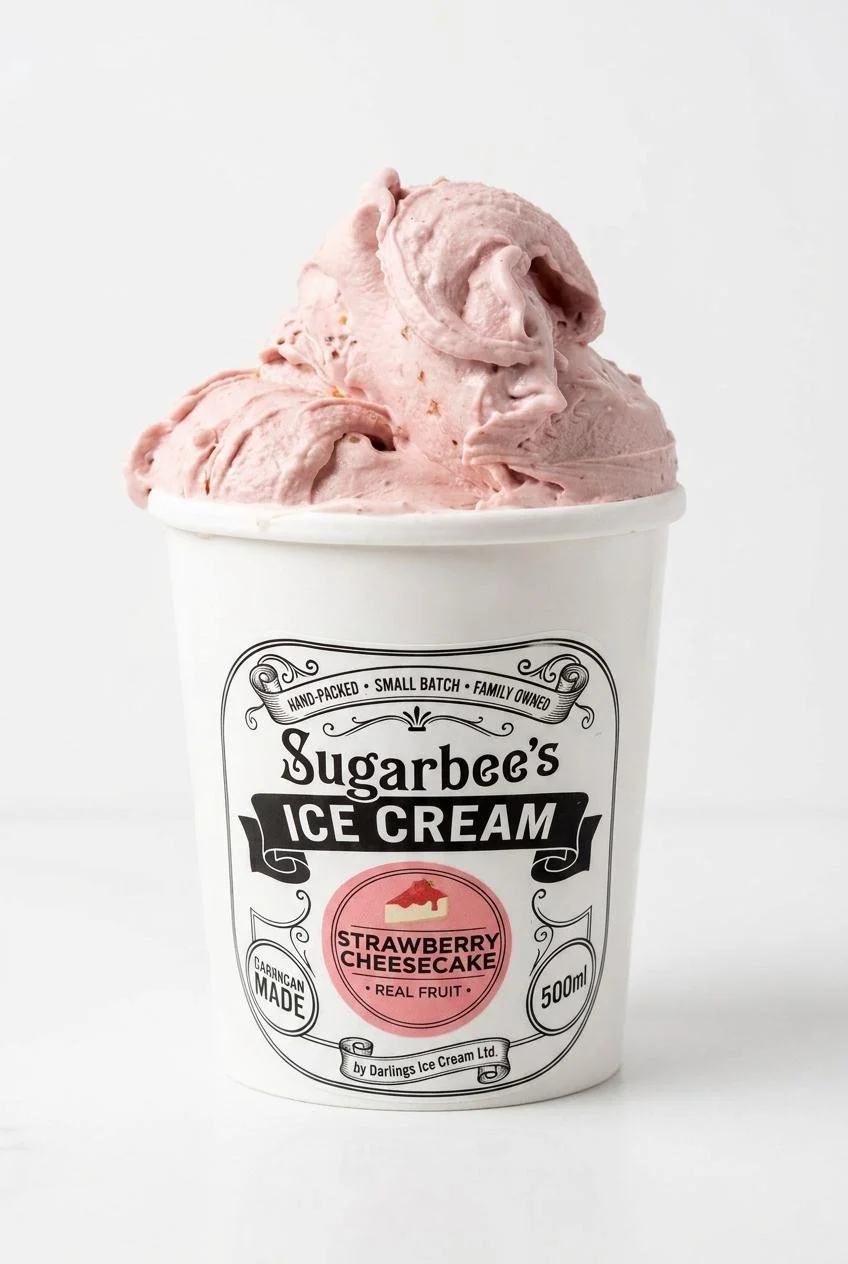 A cup of pink strawberry cheesecake flavored ice cream with a smooth, swirled texture in a white container labeled 'Sugarbee's Ice Cream, Strawberry Cheesecake, Real Fruit'. The label also states it is hand-packed, small batch, family owned, and contains 500ml.