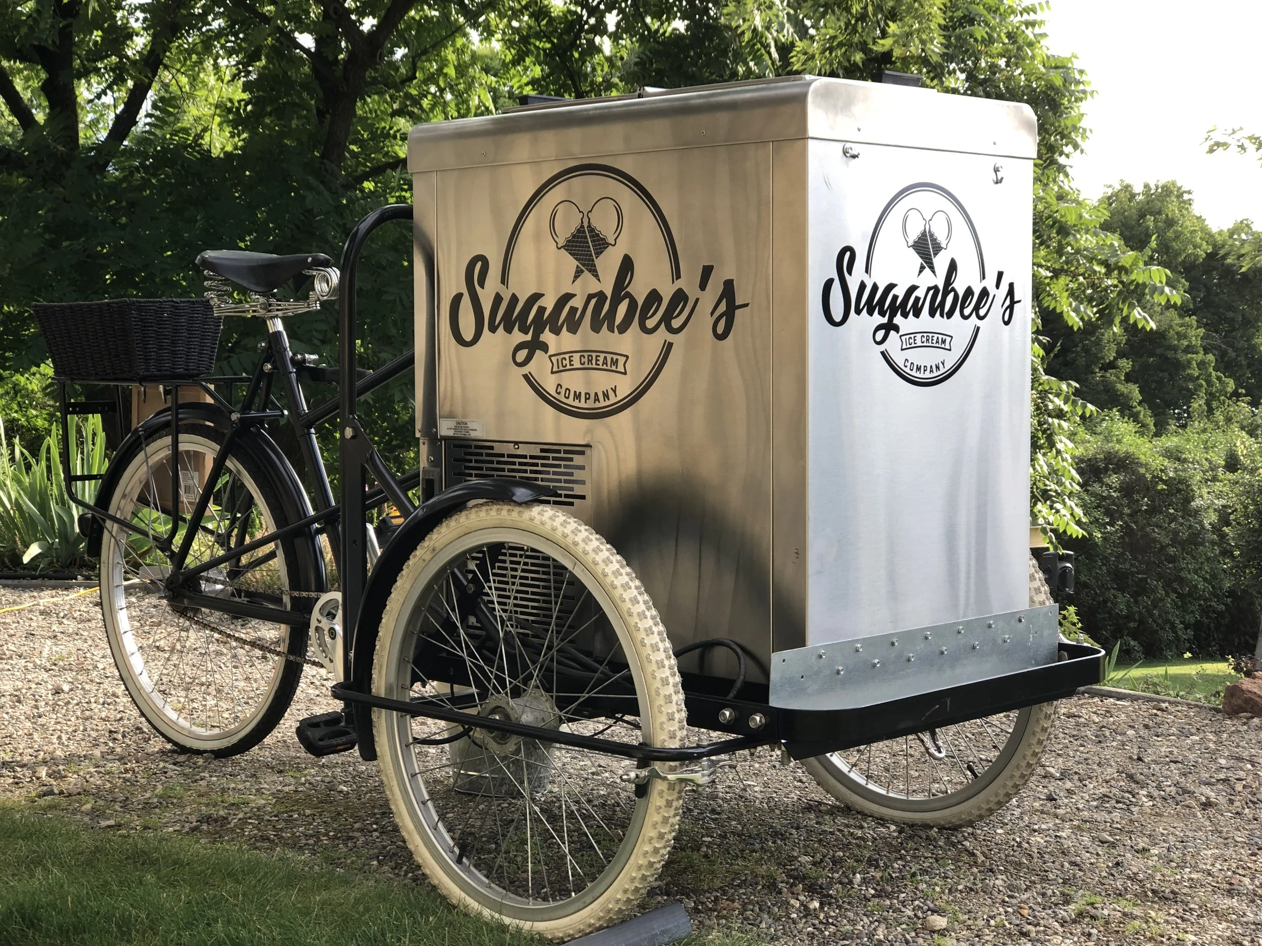 A mobile ice cream stand with the logo 'Sugarbee's Ice Cream Company,' featuring a drawing of two ice cream cones with bees on top, attached to a bicycle with a basket in front, set outdoors among green trees and bushes.