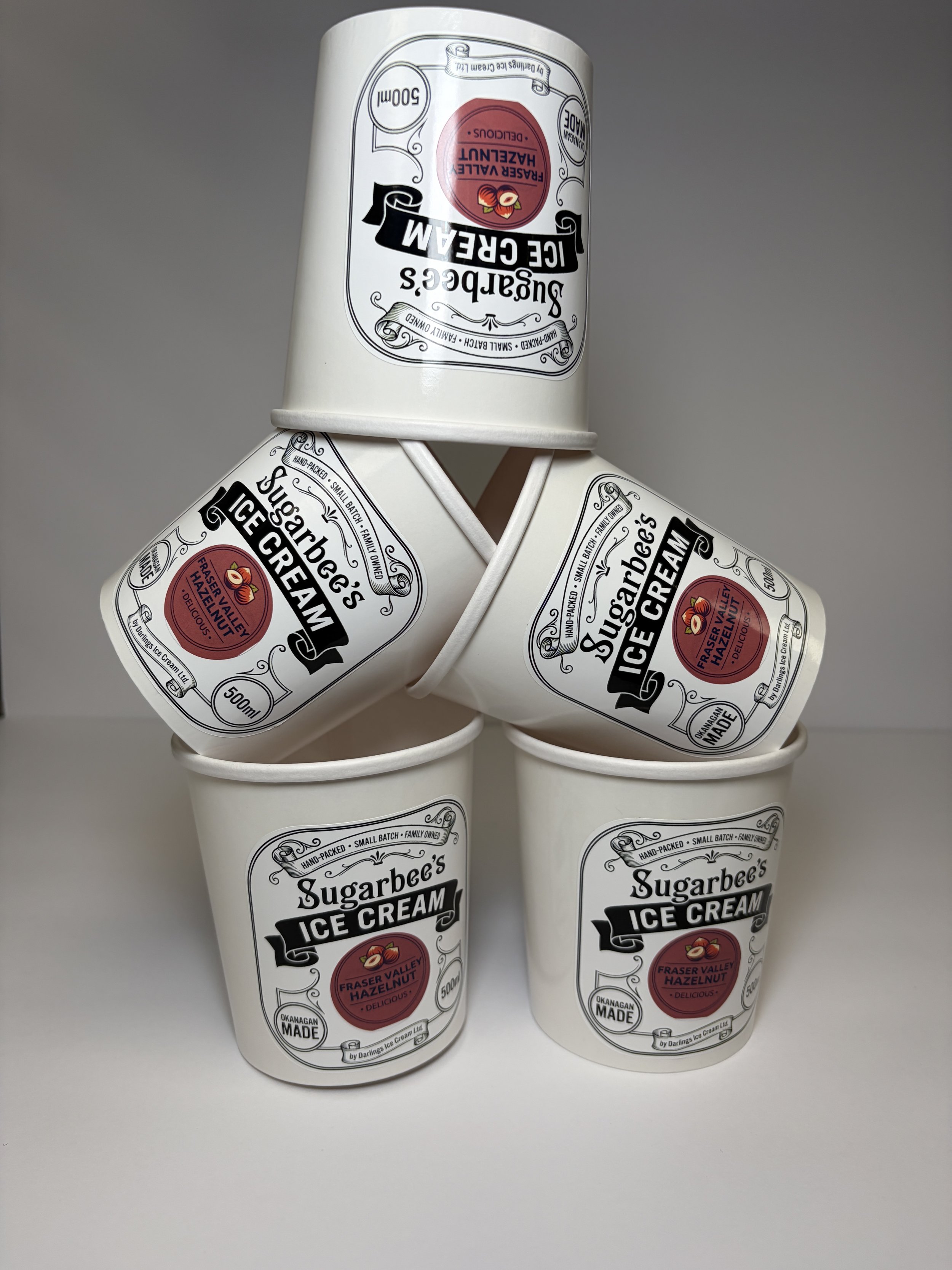 Six white cups of ice cream stacked in a pyramid formation against a gray background, with black and red labels that read 'Sugarbee's Ice Cream, Fraser Valley Hazelnut, 500ml.'
