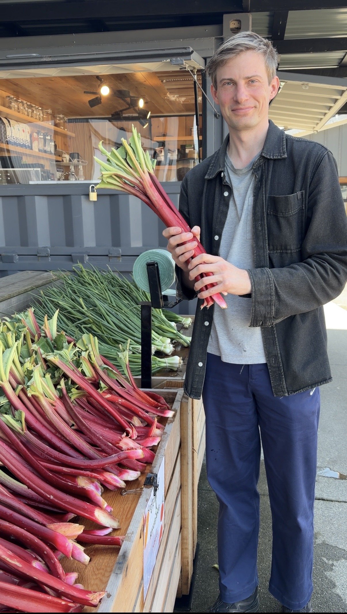 A man standing outdoors at a vegetable stand, holding a bunch of fresh rhubarb and smiling. The stand displays more rhubarb in a wooden bin, with a store window and metal roof in the background.