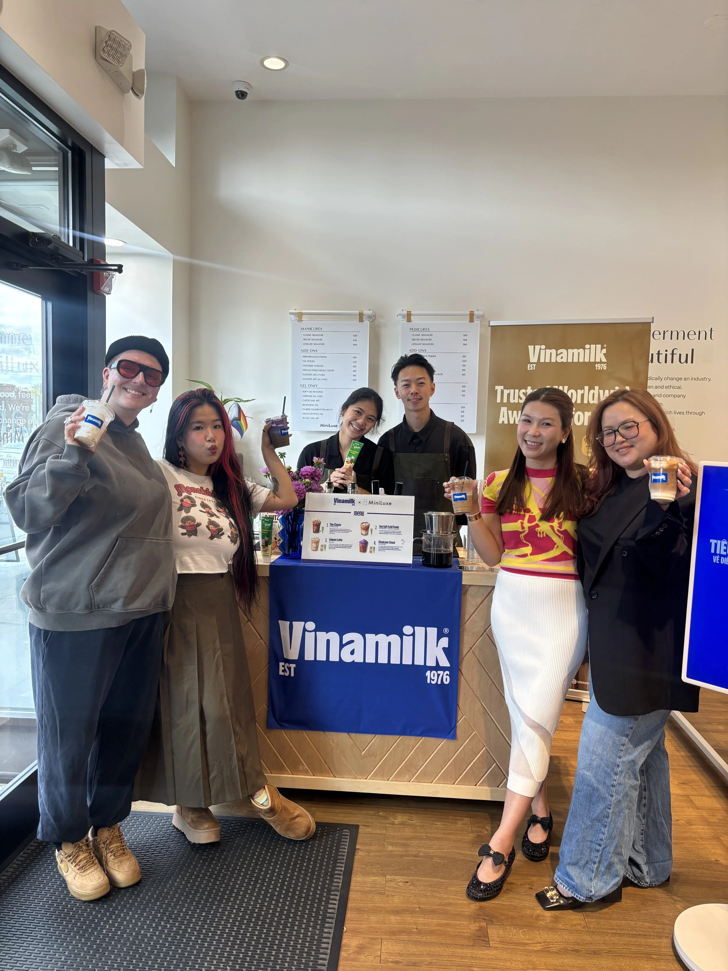 Group of six people standing behind a Vinamilk counter inside a store, holding up drinks and smiling at the camera.