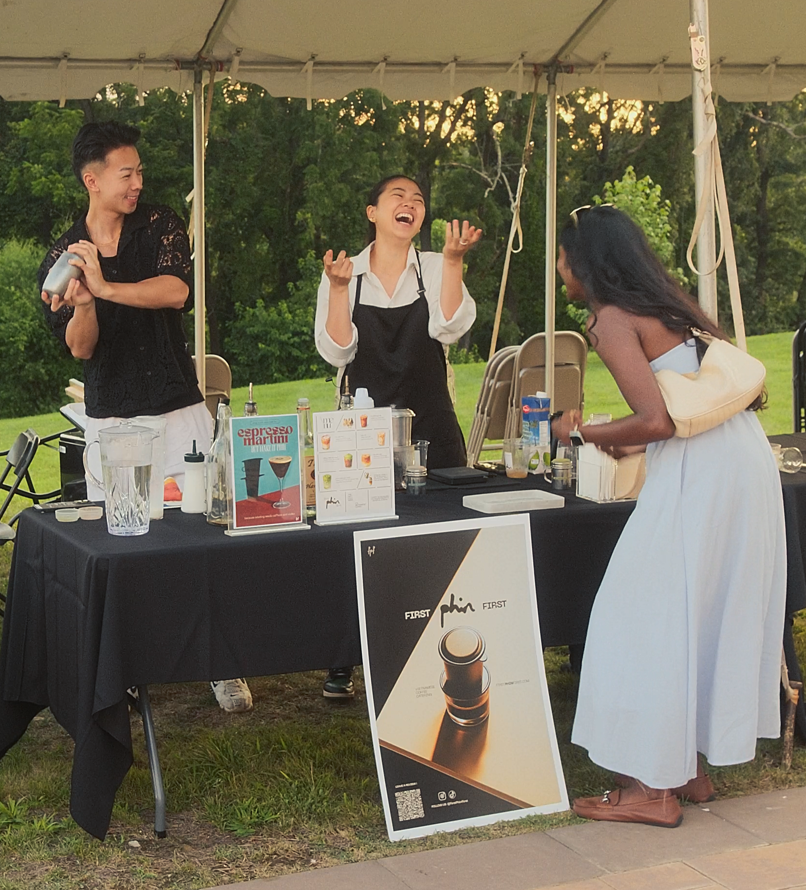 Three women at an outdoor booth laughing and talking. One woman is working behind the booth with items and a menu, another woman is standing in front, smiling, and the third woman is leaning over the table.