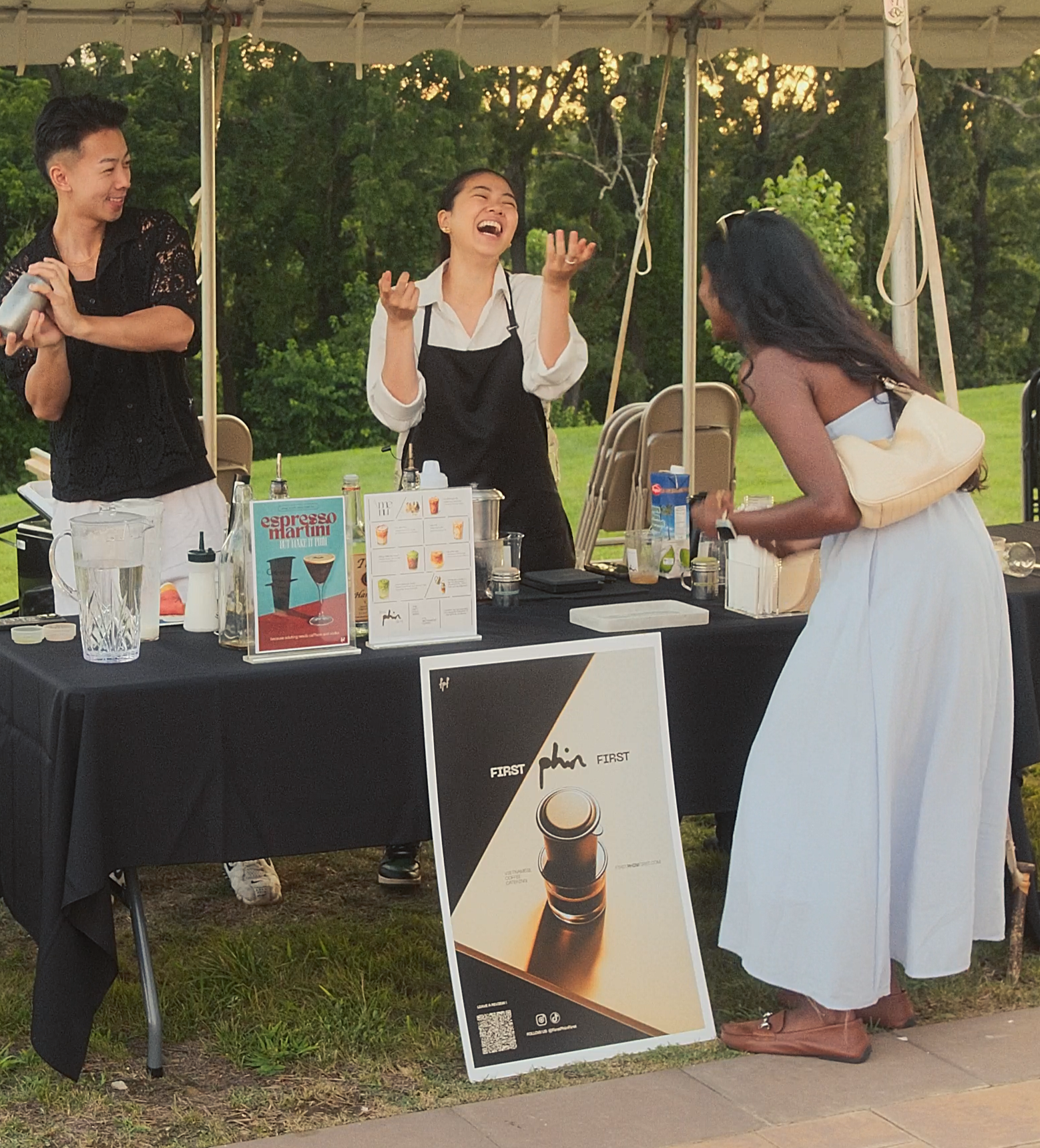 Three people at an outdoor coffee stand laughing and talking, with a woman in a white dress and a butterfly hair clip in front of the stand, and two women behind the counter, one wearing a black apron and the other holding a cup.