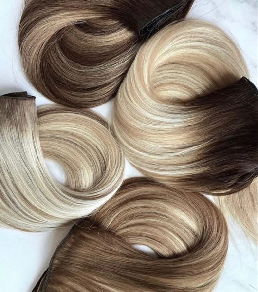 Assorted hair extensions in shades of brown, blonde, and black, arranged in loose waves on a white background.