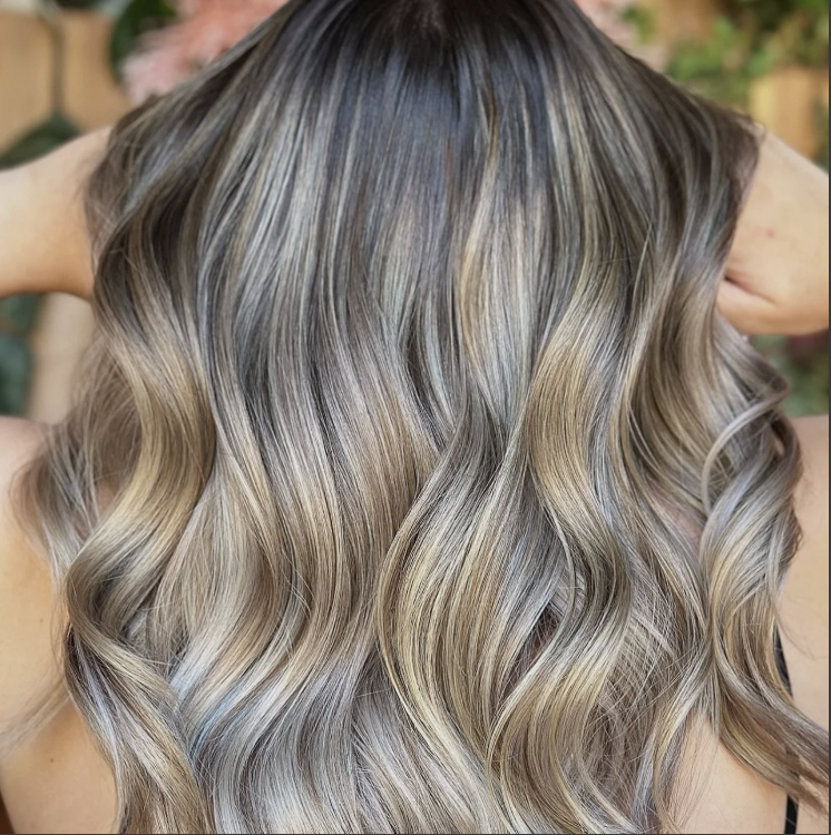 A person with long, wavy blonde and light brown hair seen from the back, with a blurred background of plants.