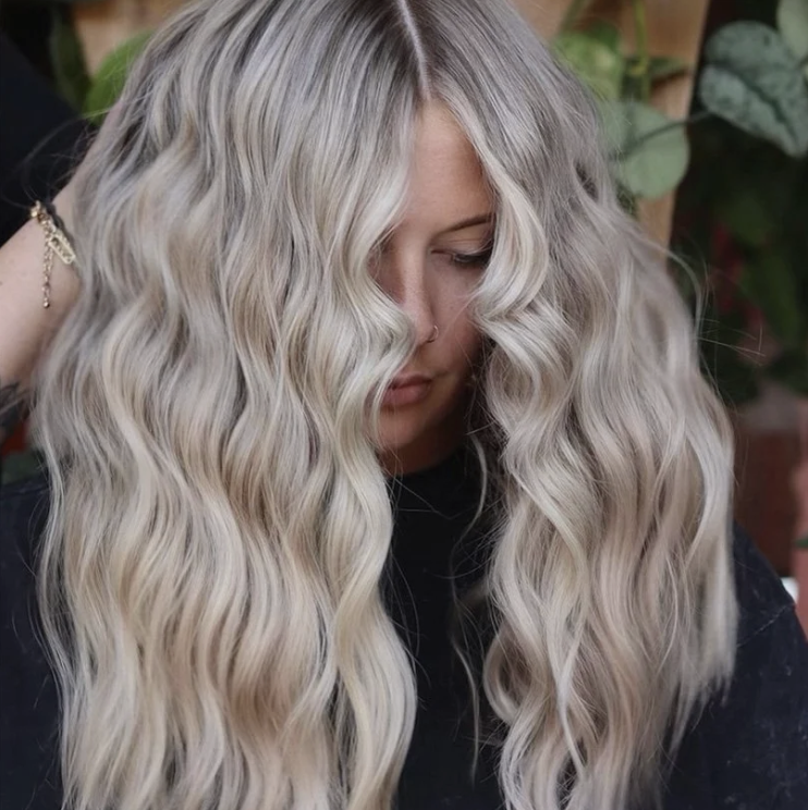 A woman with long, wavy platinum blonde hair with darker roots, looking down with part of her face obscured.