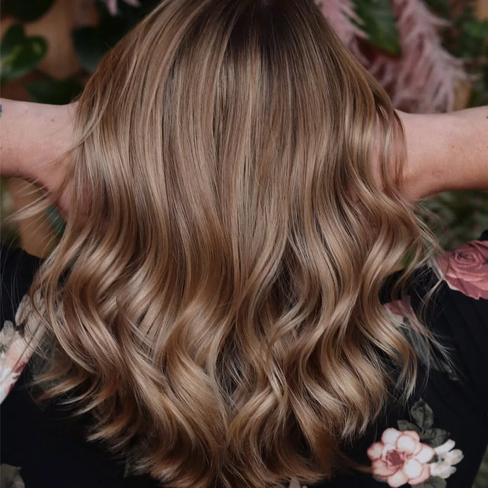 Woman with wavy, shoulder-length blonde hair, caught from behind with hands behind her head, surrounded by green and pink foliage.