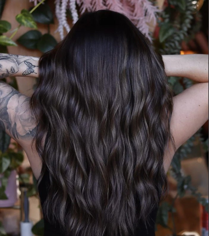 Back view of a woman with long, wavy dark brown hair, standing in front of leafy plants.