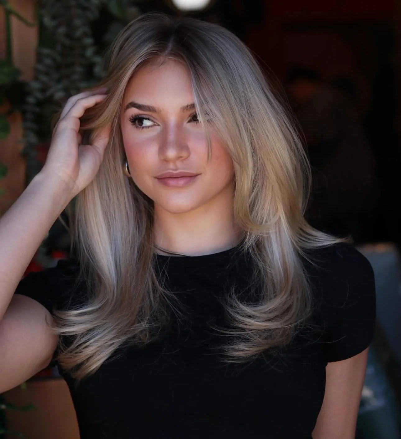 A young woman with blonde hair and hoop earrings, wearing a black top, looks slightly to her left with her hand touching her hair.
