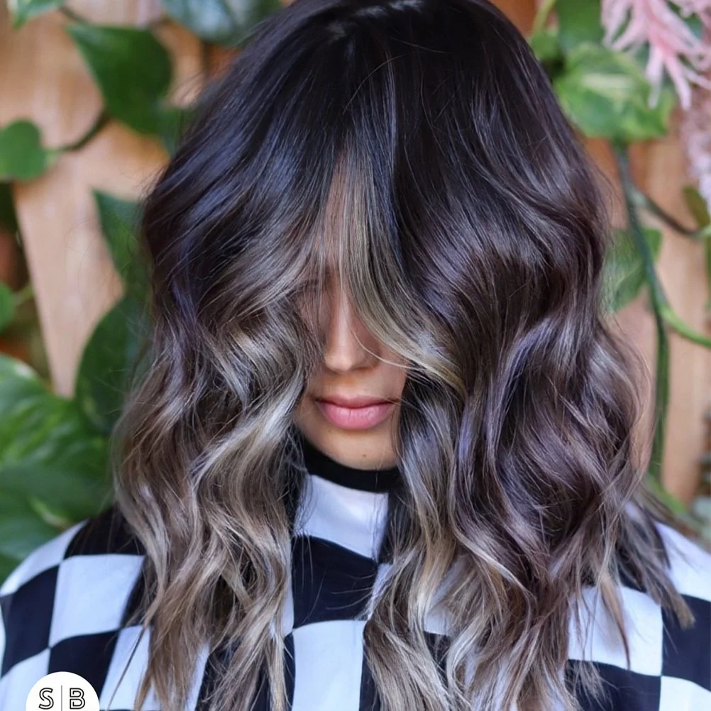 A woman with wavy, shoulder-length hair styled in loose curls with dark brown roots transitioning to blonde highlights, partly covering her face, standing in front of green leafy plants.