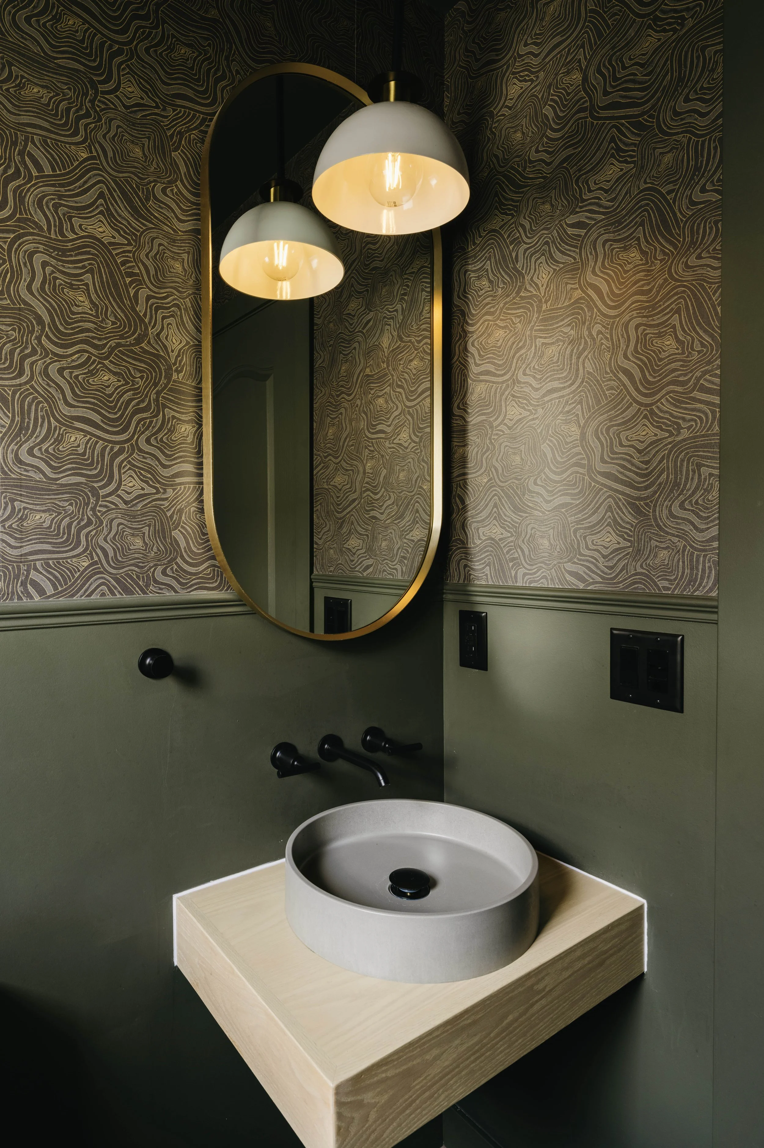 Modern bathroom with a round stone sink on a light wood countertop, a large mirror with an oval brass frame, two white ceiling pendant lights, dark wall outlets, and patterned wallpaper.