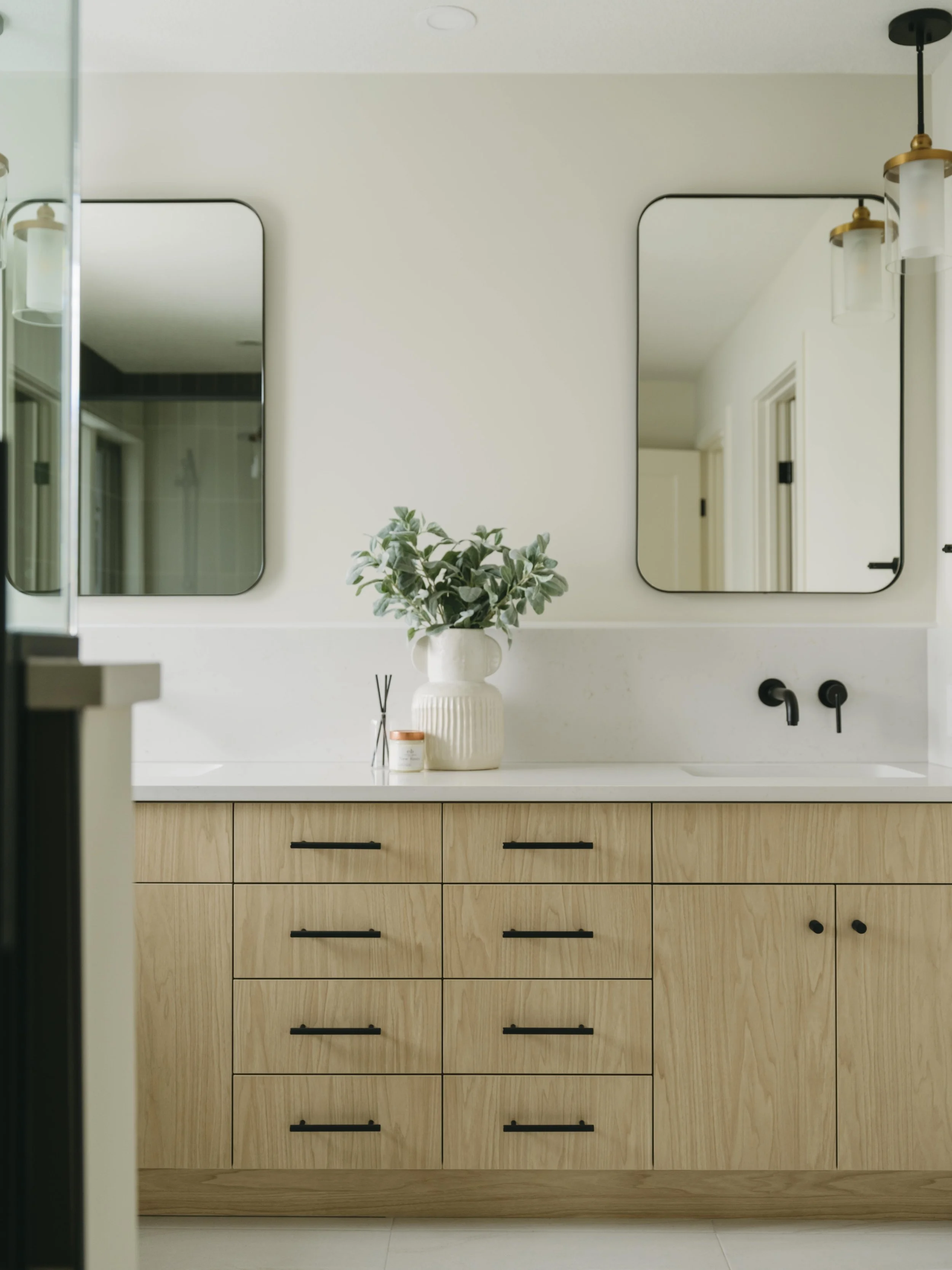 Modern bathroom vanity with wooden drawers, black handles, a white countertop, a large white vase with green foliage, and two square mirrors with black trim above the sink. There are two black faucets and a light fixture with glass shades on the wall.