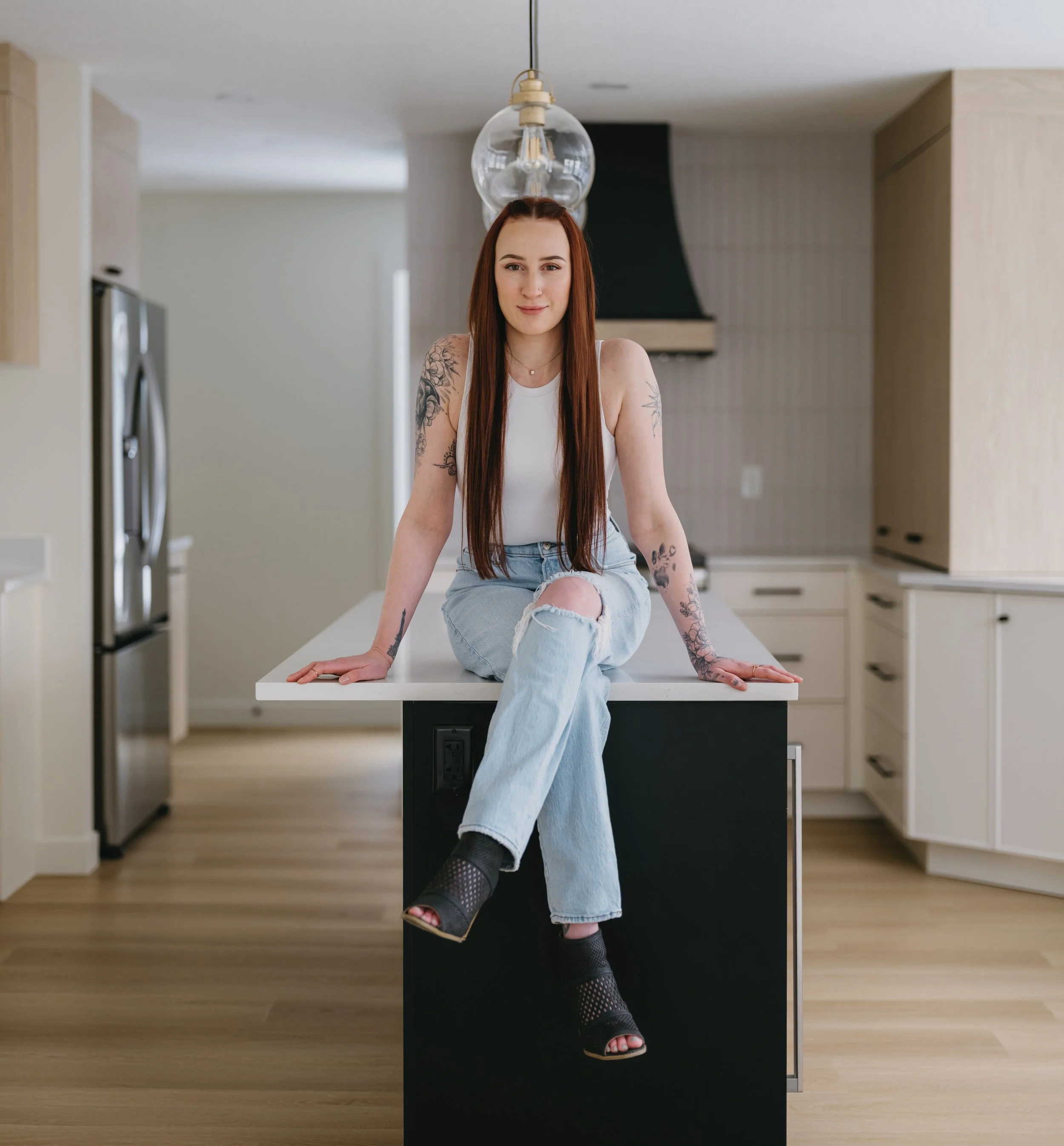 A young woman with long red hair and tattoos sitting on a kitchen island in a modern kitchen with light wood cabinets and a black range hood.