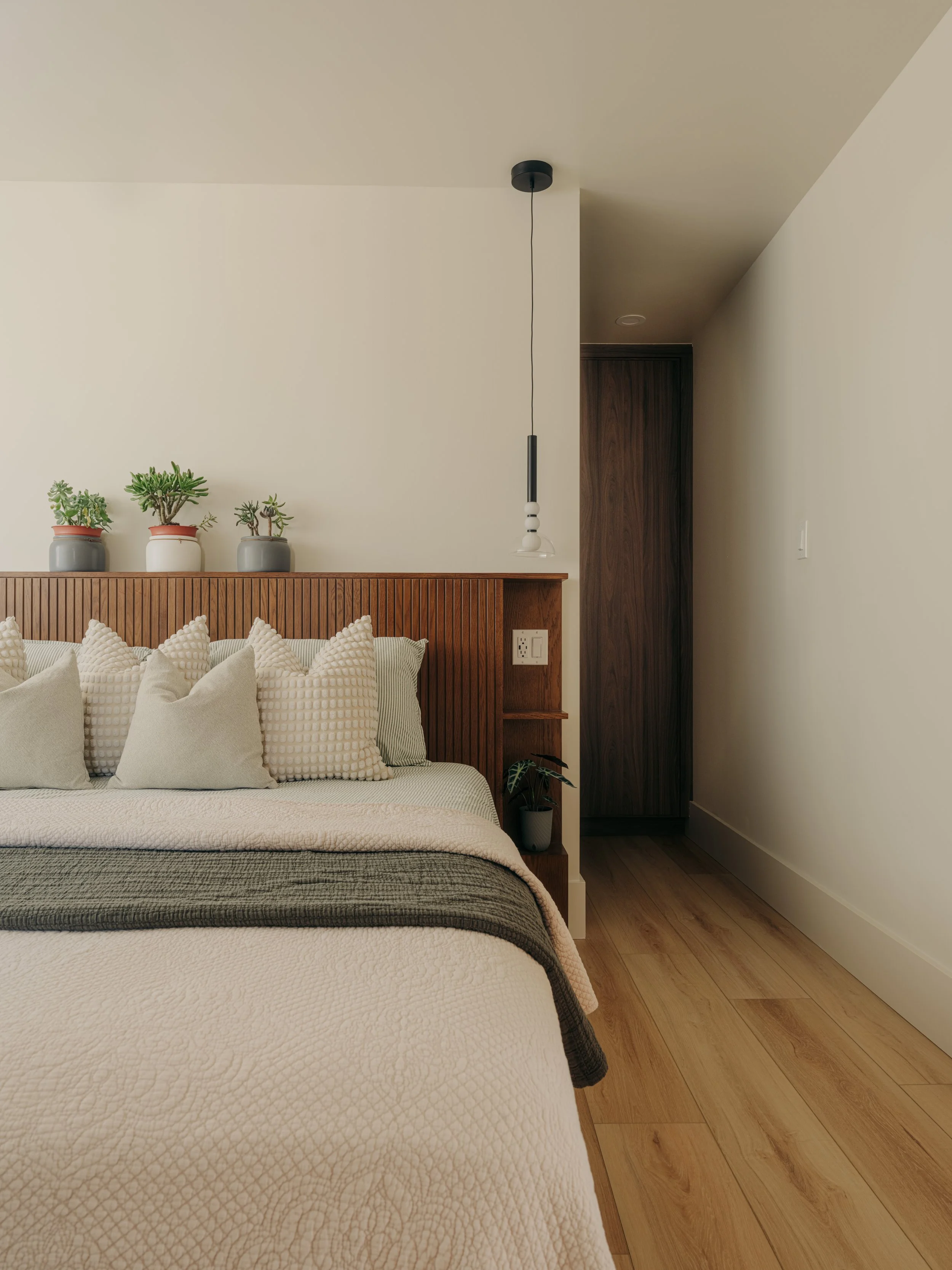 A cozy bedroom with a bed made with white textured bedding and several white pillows. There is a wooden headboard with a built-in shelf holding three potted succulent plants. A minimalist pendant light hangs above the headboard. The room has light-colored wooden flooring and neutral-colored walls.