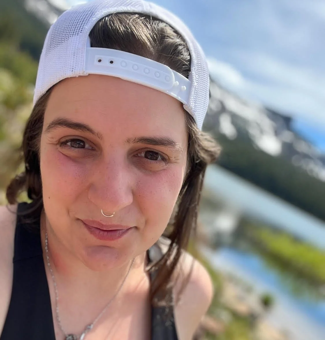 A nonbinary person smiling outdoors near a body of water with snow-capped mountains in the background, wearing a white baseball cap backwards and a black tank top.