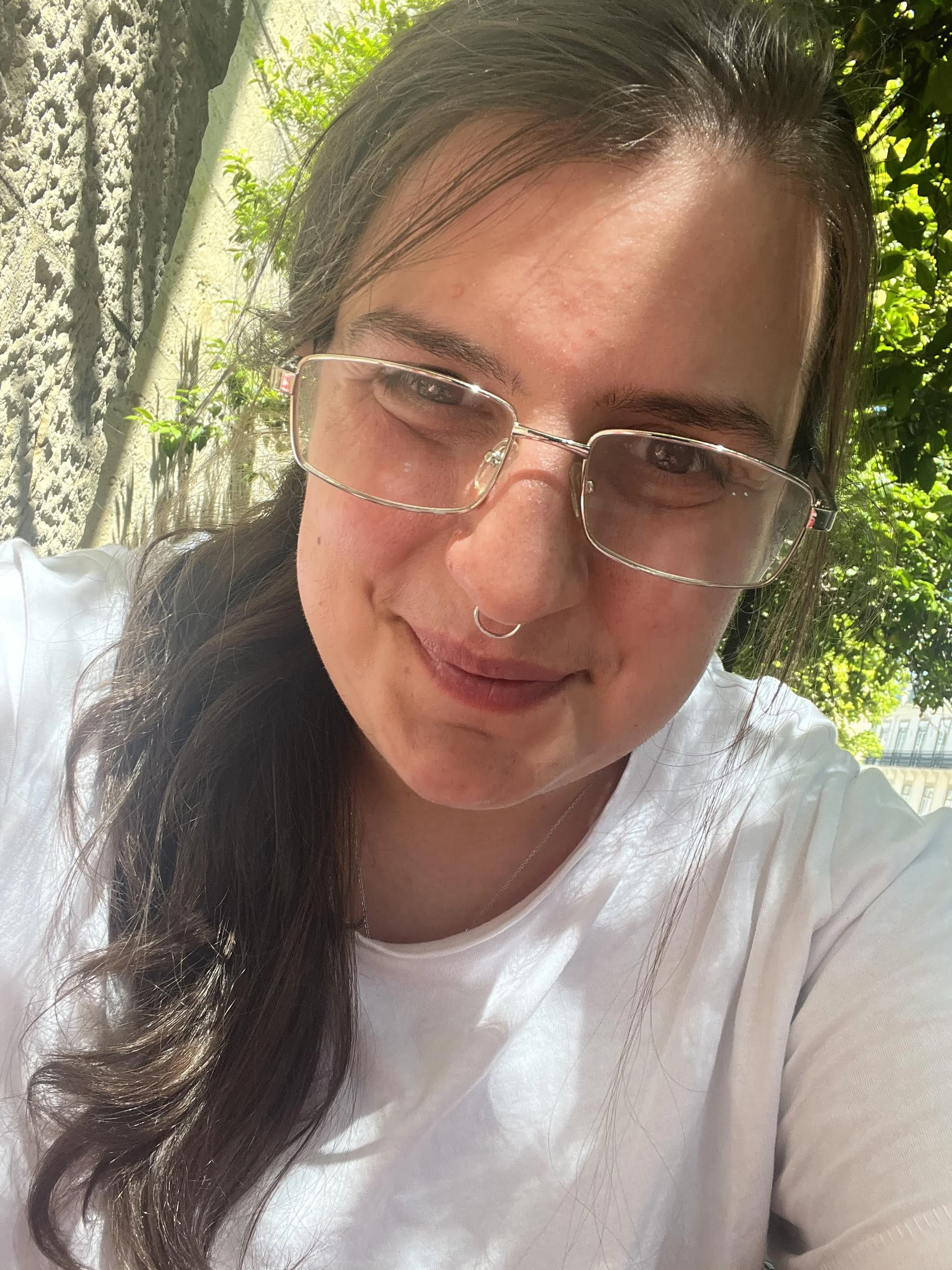 A person with glasses and a septum piercing smiling outdoors on a sunny day, with green foliage and a textured stone wall in the background.