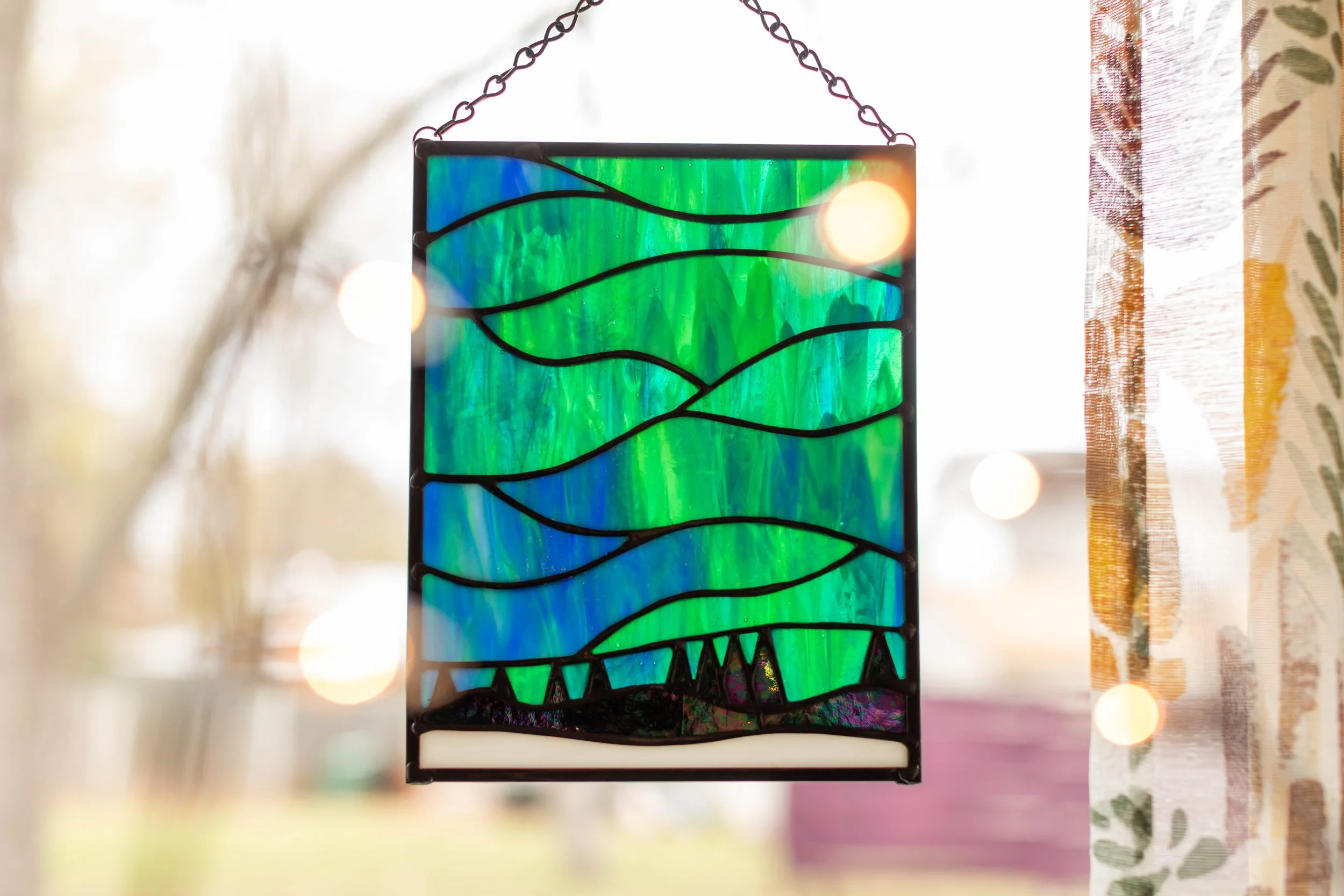 Colorful stained glass hanging window with northern lights and treescape, hanging near a window with blinds.