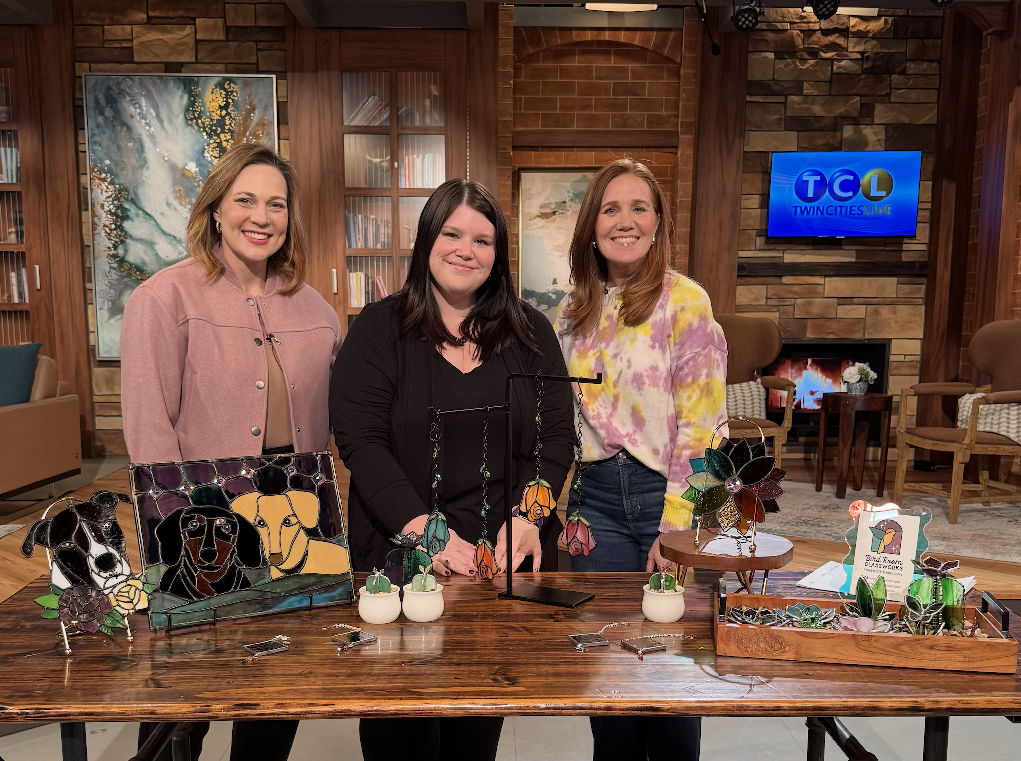 Three women standing behind a wooden table displaying stained glass art in a cozy TV studio setting, with a TV screen showing 'TCL Twincities Live' in the background.