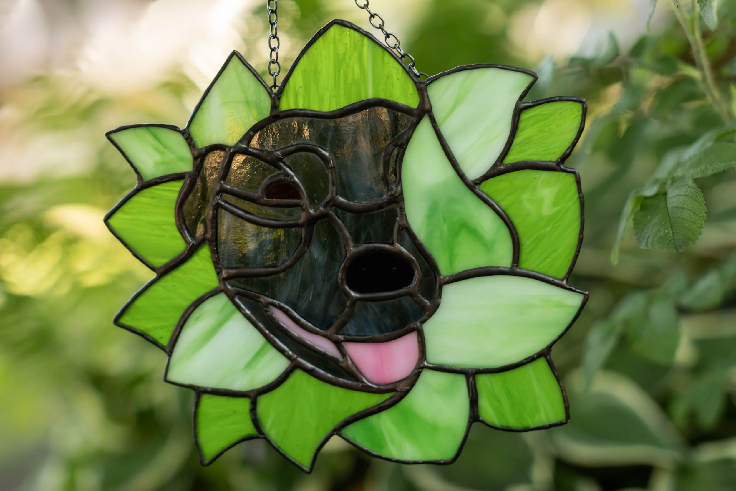 Stained glass garden decoration shaped like a puppy face with green petals surrounding it, hanging on a chain among green foliage.