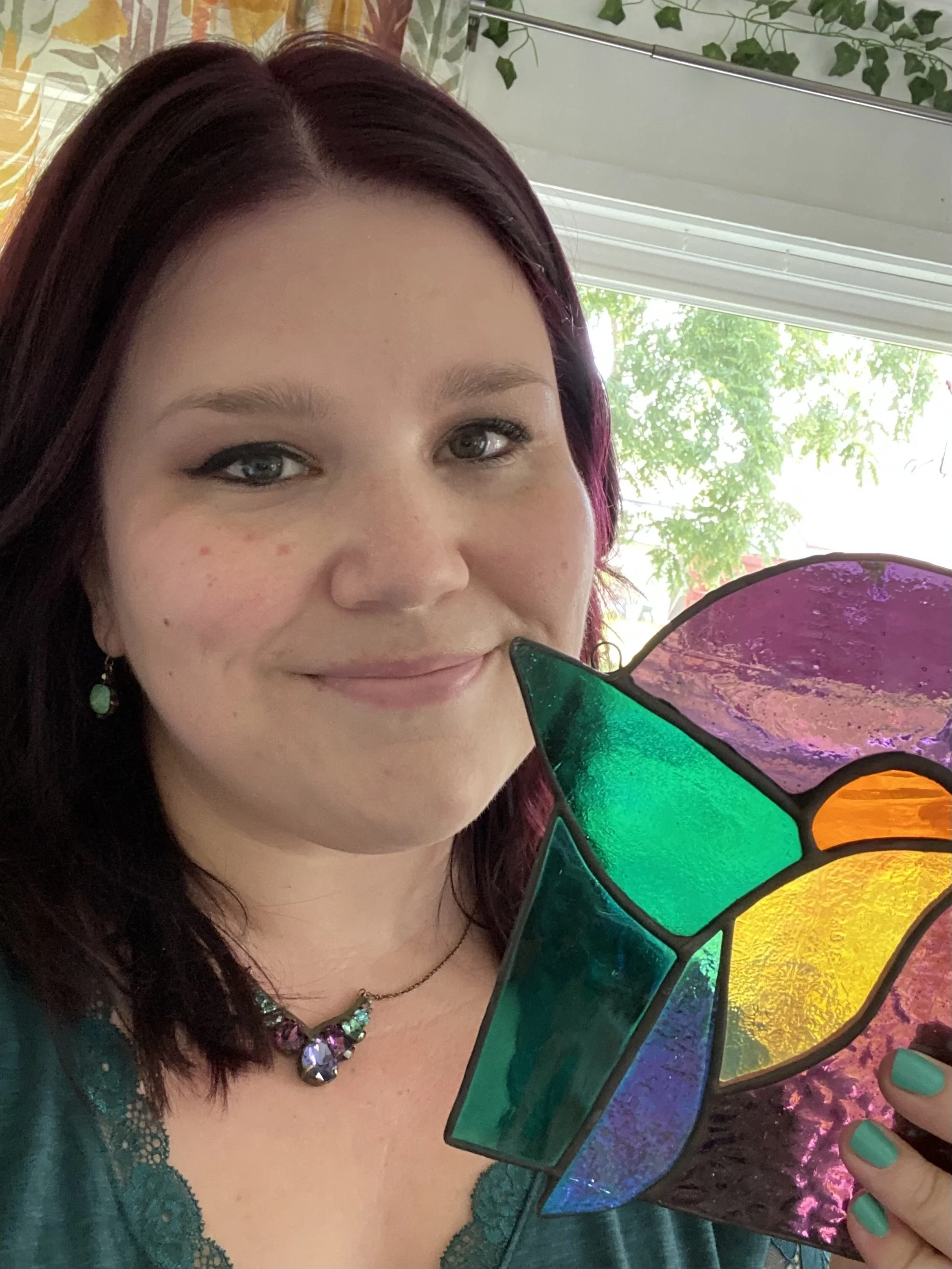 A woman with shoulder-length dark purple hair, wearing green earrings and a necklace with purple gems, holding a colorful stained glass piece resembling a flower or butterfly. She is smiling and standing near a window with pink, yellow, green, purple, and blue glass sections, with trees visible outside.