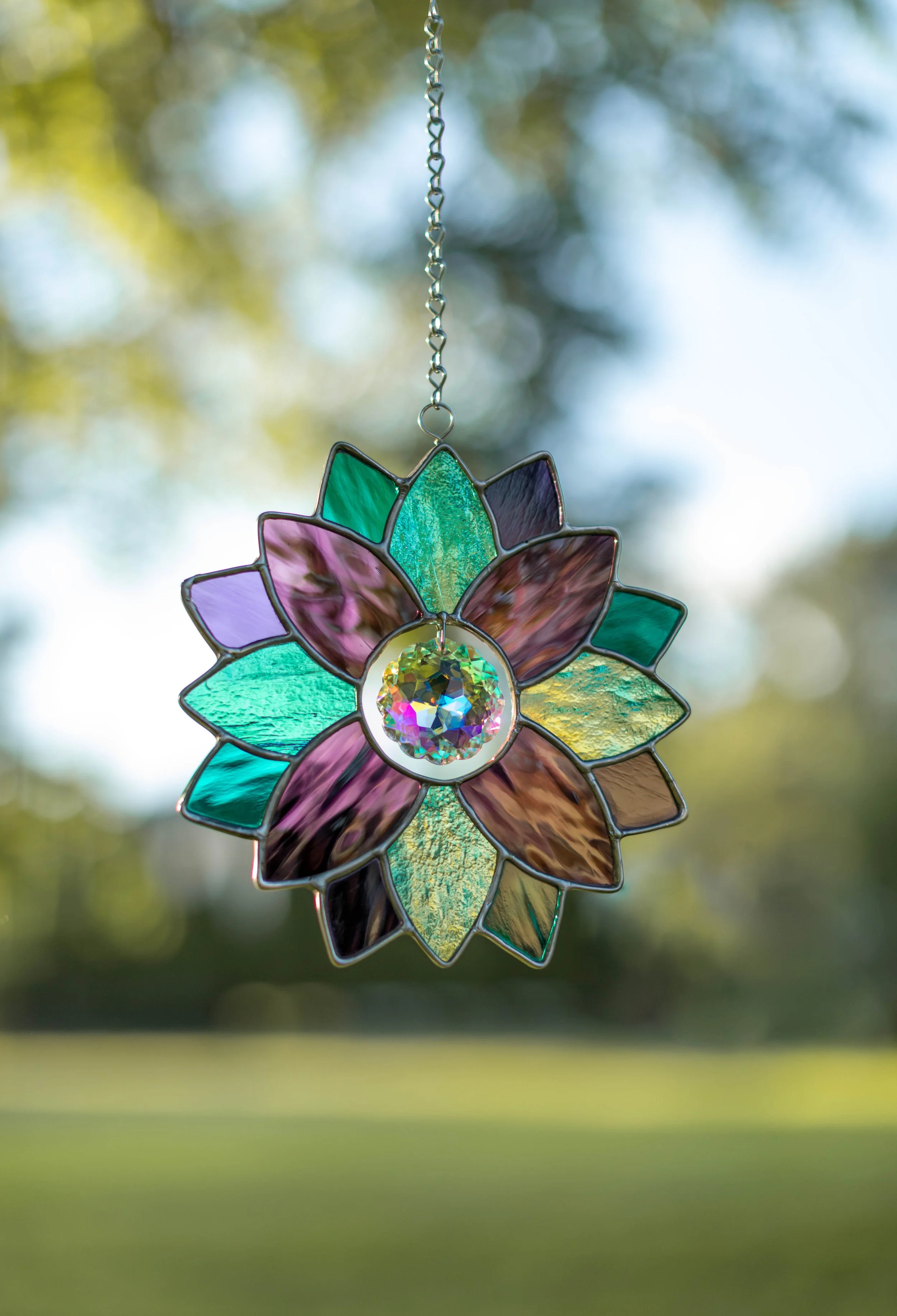 A stained glass floral ornament hanging outdoors with blurred trees and sky in the background.