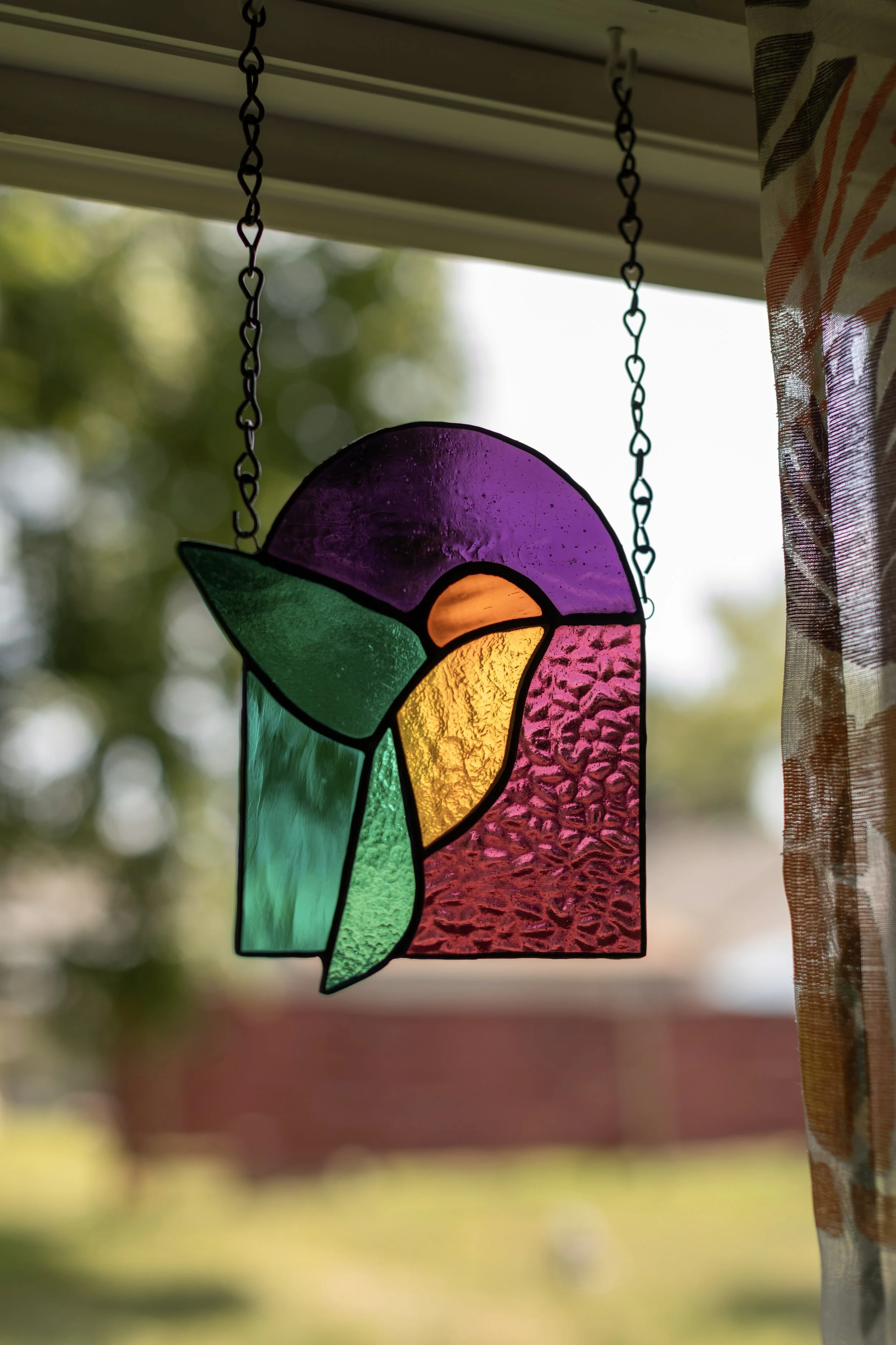 Colorful stained glass hummingbird decoration hanging in front of a window with a blurred outdoor background.