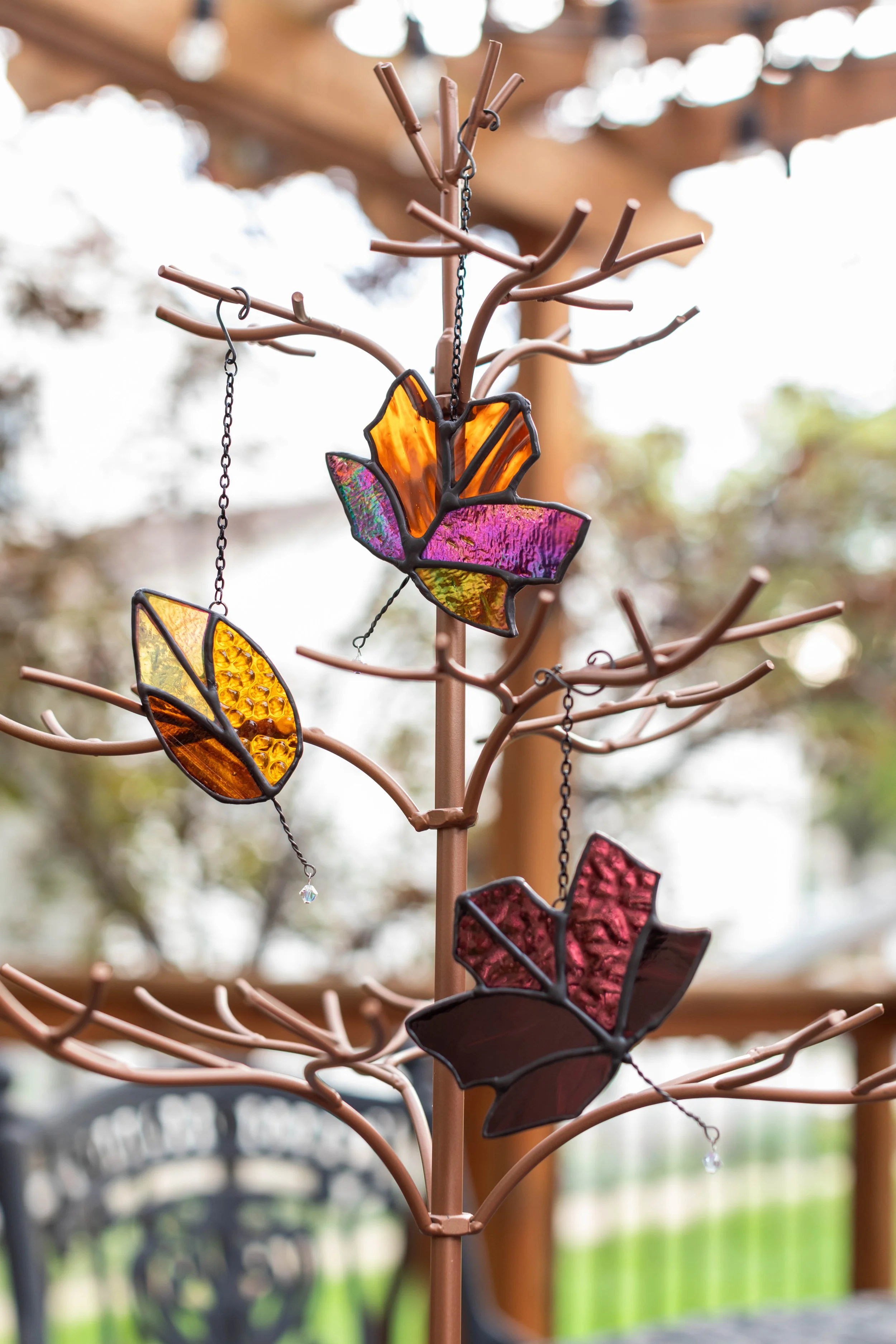 A decorative metal tree with stained glass leaf embellishments hanging from its branches, outdoors with a blurred background.