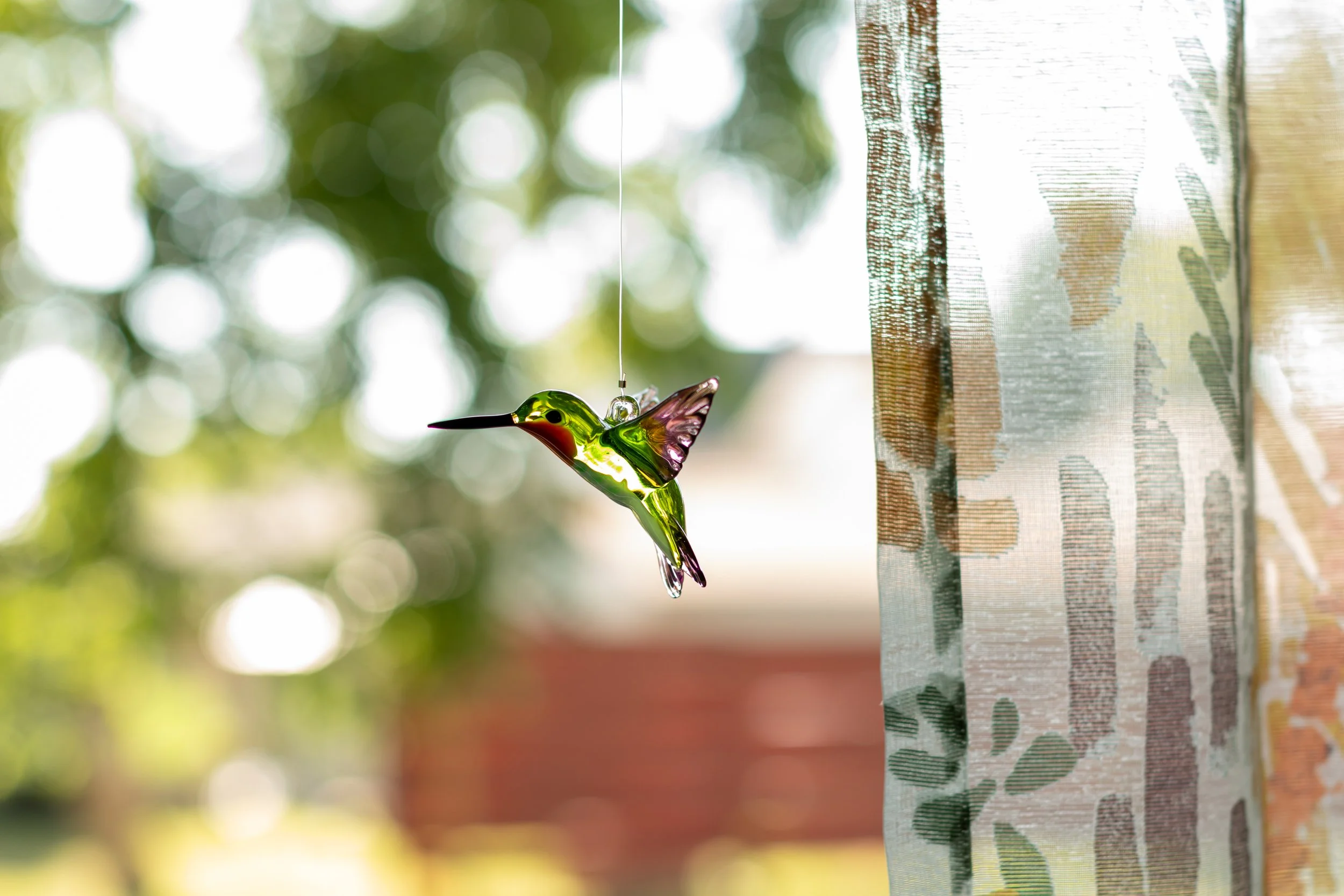 A glass hummingbird decoration hanging in front of a window with blurred greenery outside and a patterned curtain on the right side.