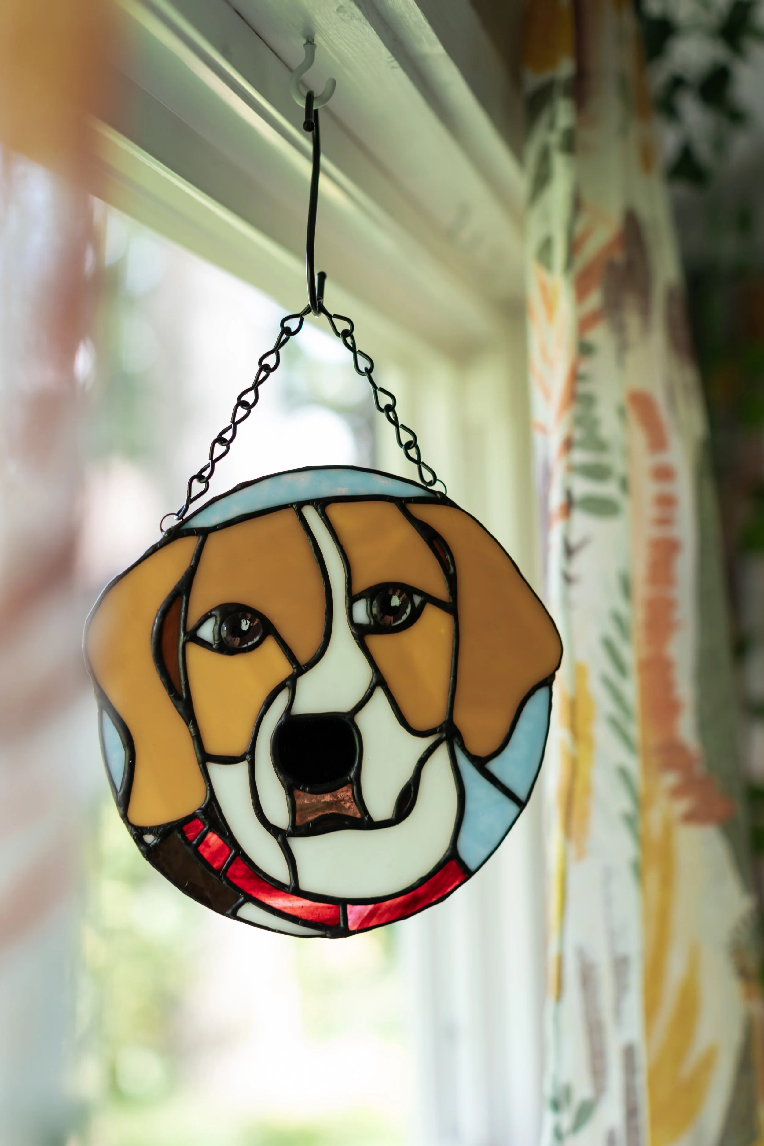 Stained glass window hanging of a dog's face with brown, white, and black colors, suspended by a chain from the ceiling near a window.