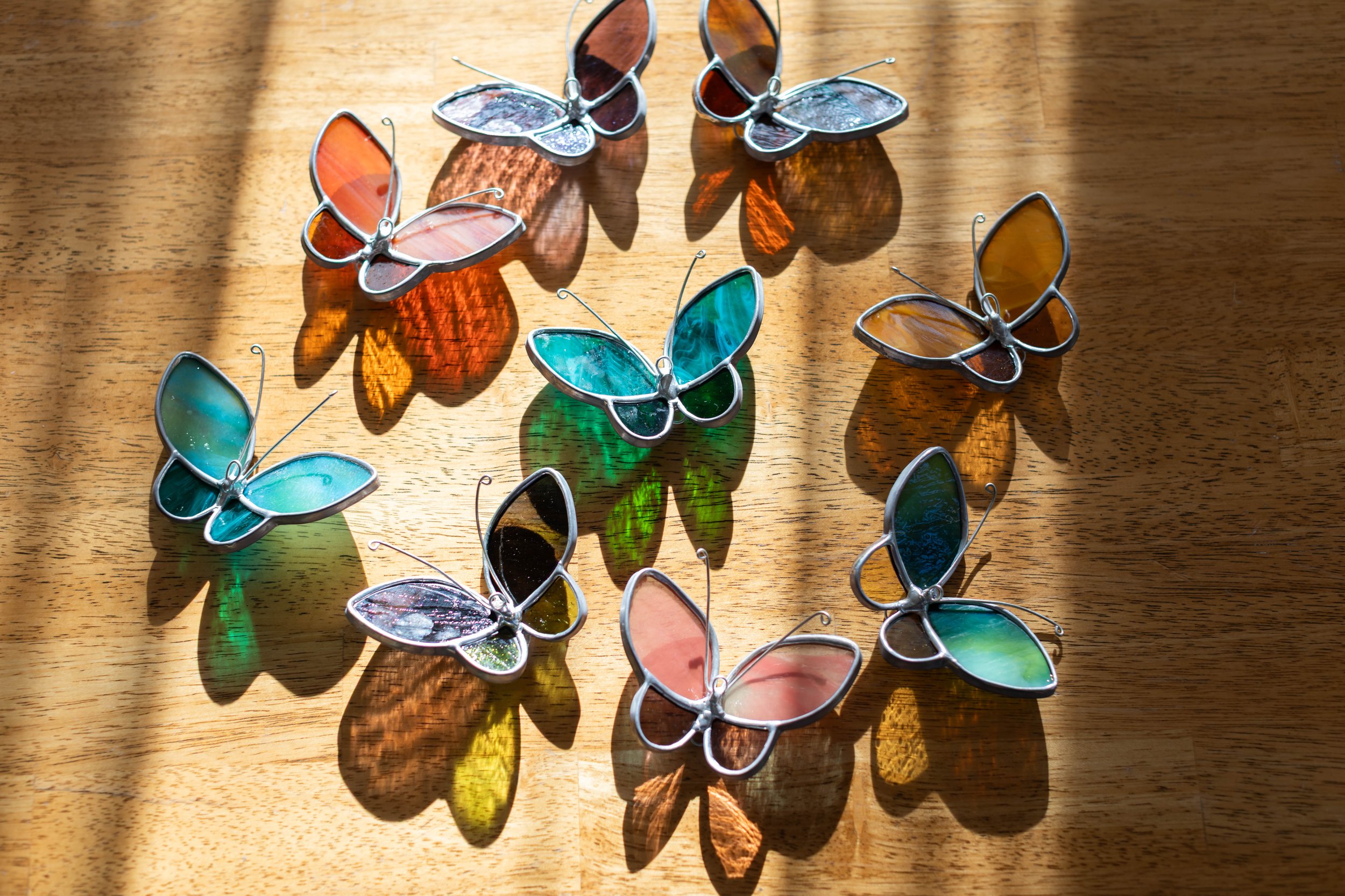 Colorful stained glass butterfly ornaments arranged on a wooden surface with sunlight casting colorful shadows.