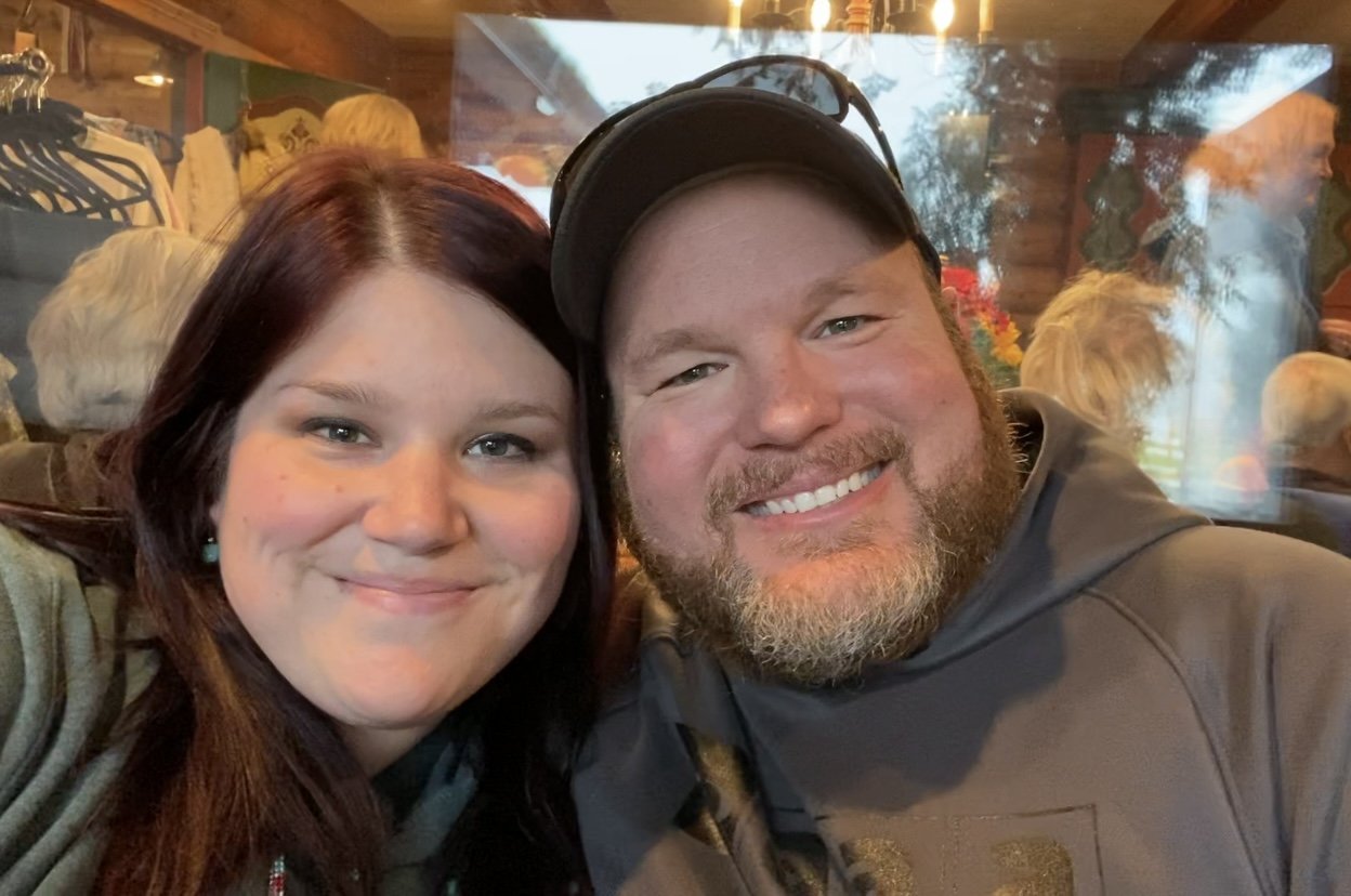 A smiling woman with long dark hair and a man with a beard and a baseball cap taking a selfie together in a cozy, rustic indoor setting with people and decorations in the background.