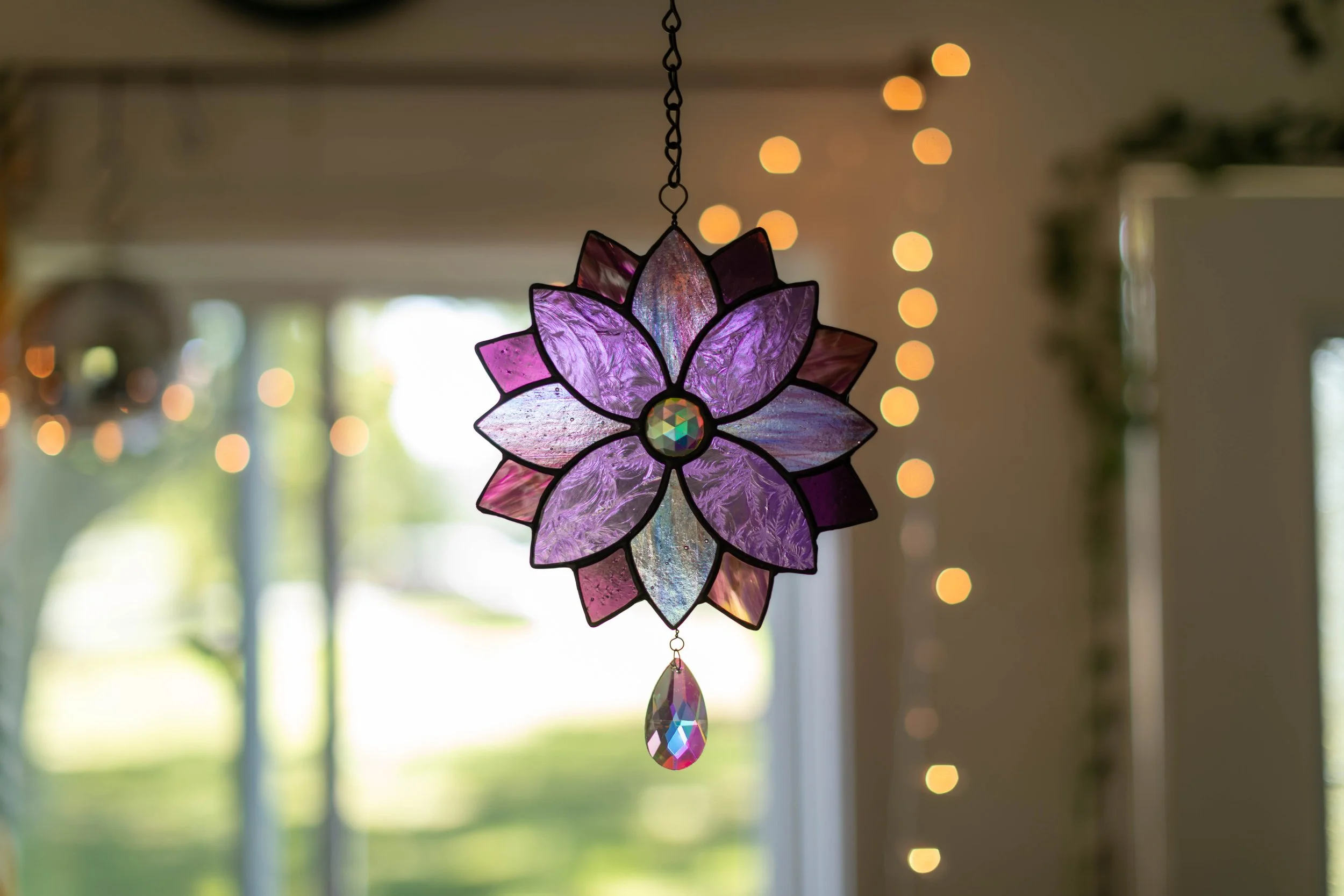 A stained glass hanging decoration in the shape of a purple and pink flower with a colorful glass gem in the center and a teardrop-shaped gem hanging below. Blurred indoor background with string lights and windows showing greenery outside.