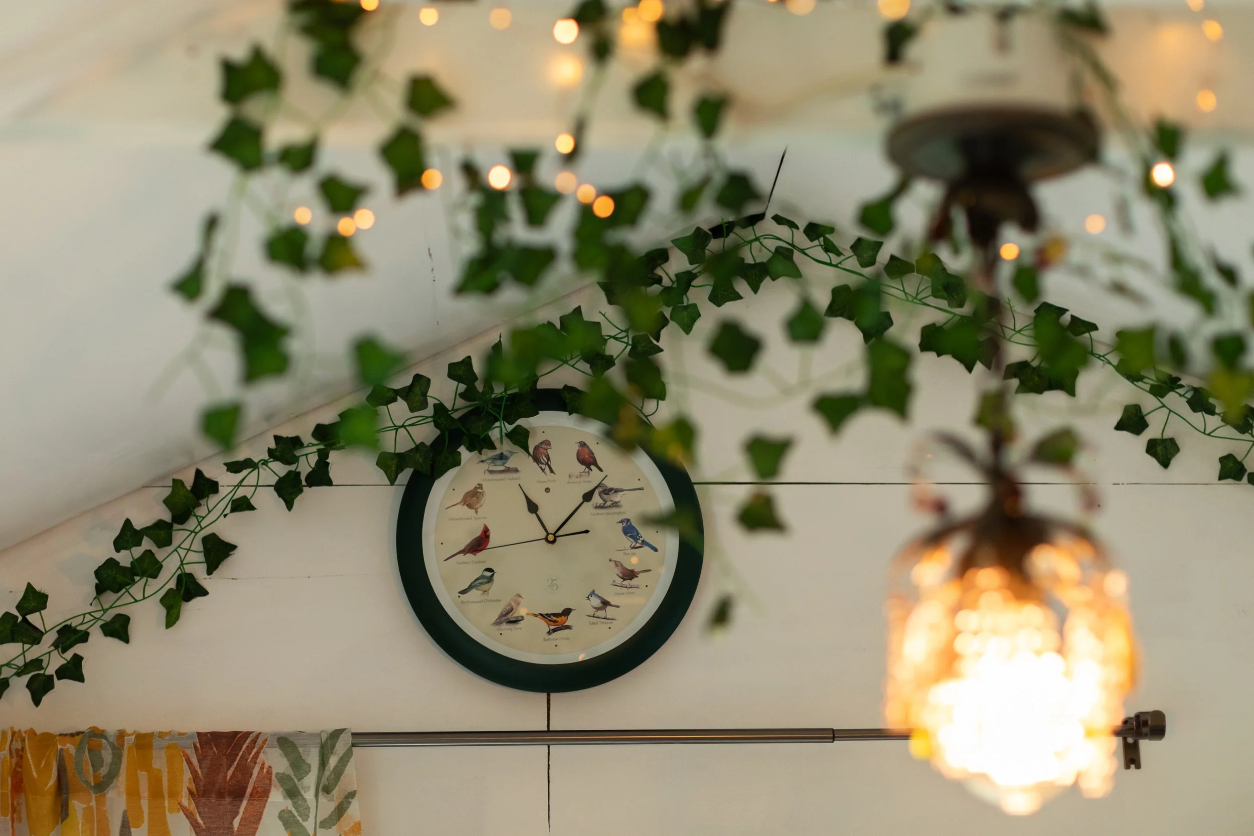 A round wall clock with bird illustrations instead of numbers, hanging on a white wall decorated with green ivy and string lights. Part of a colorful curtain is visible at the bottom, and a blurred hanging light fixture is in the foreground.
