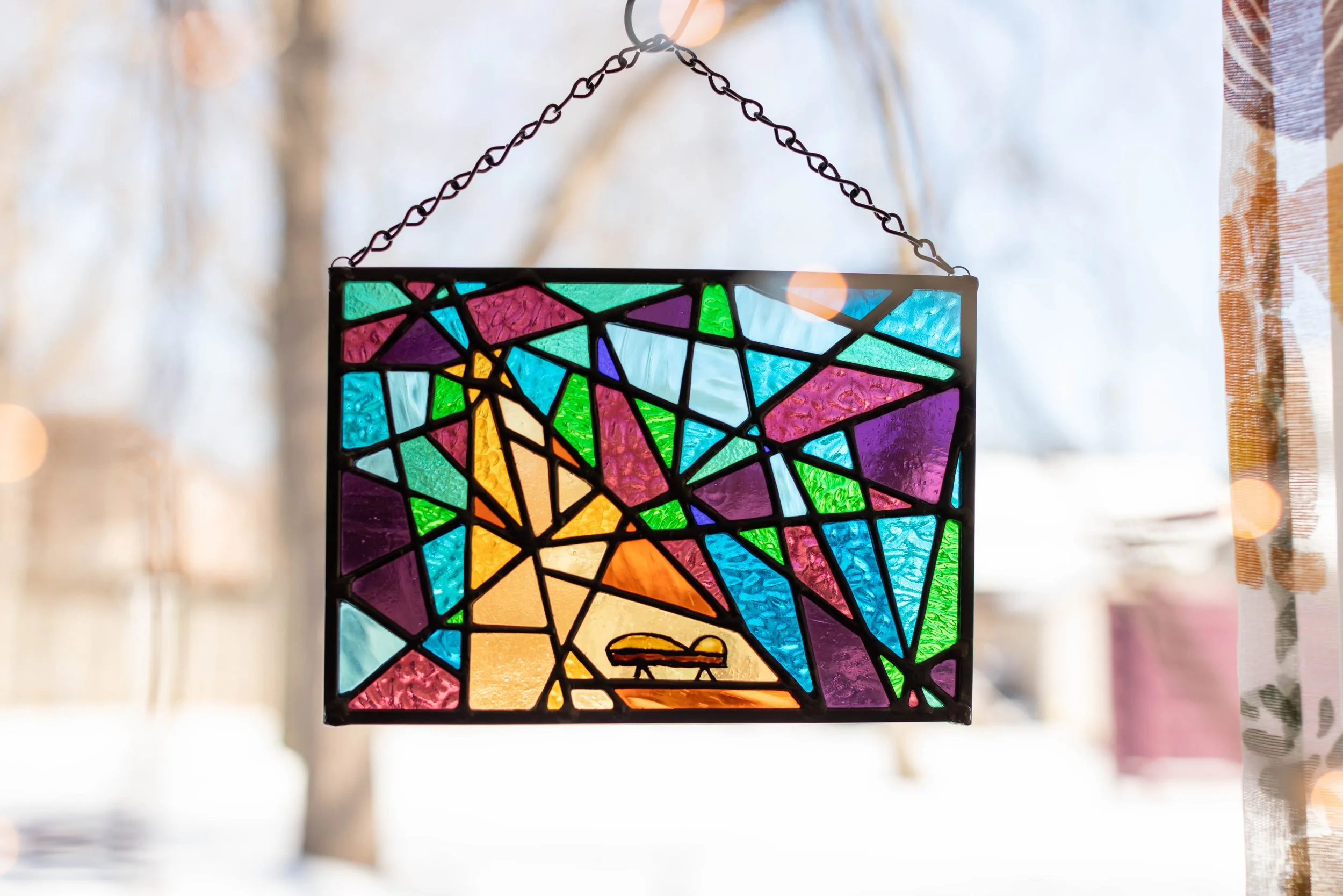 Colorful stained glass window hanging outside with blurred trees and sunlight in the background.