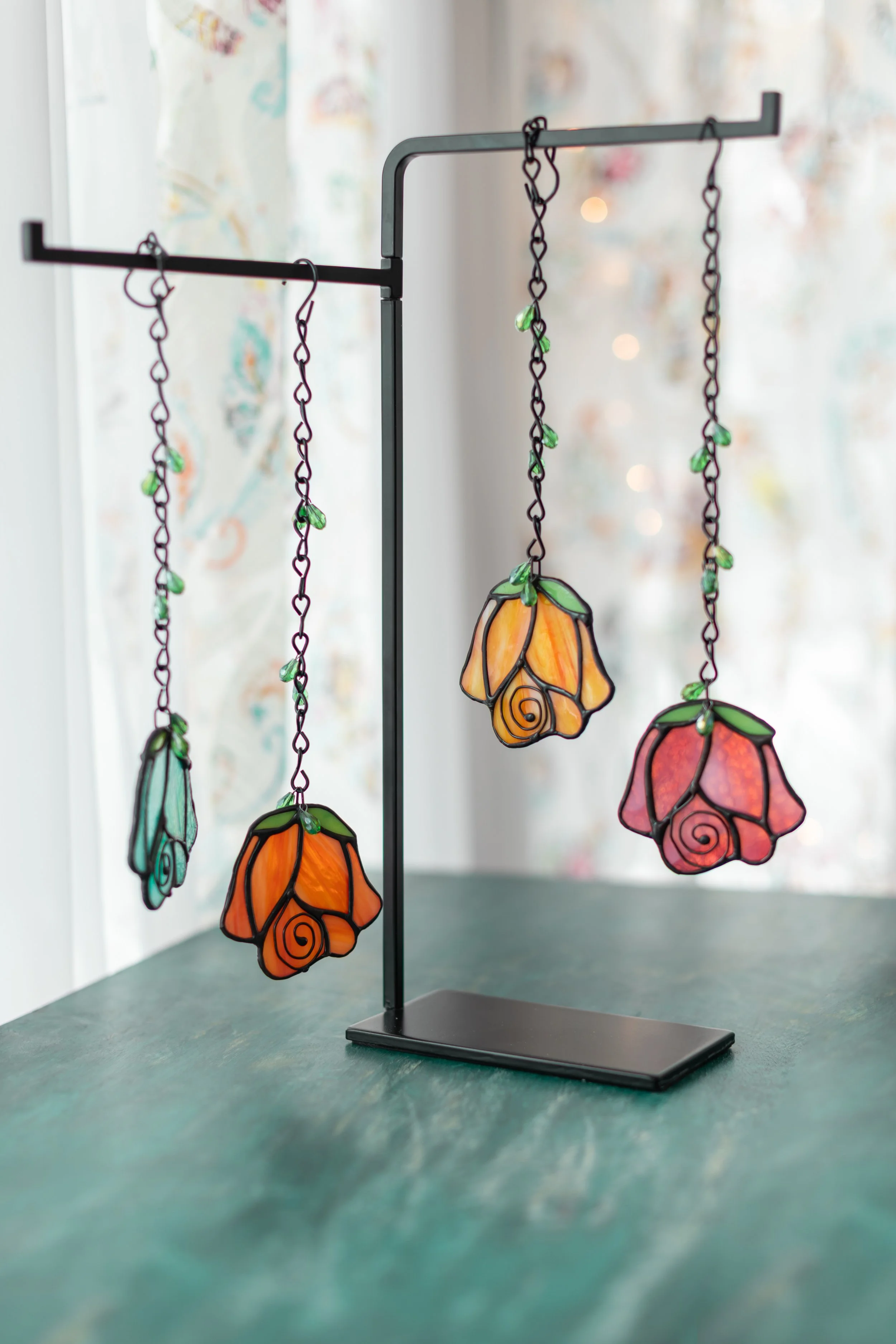Colorful stained glass flower-shaped hanging ornaments on a black metal display stand on a green table indoors.