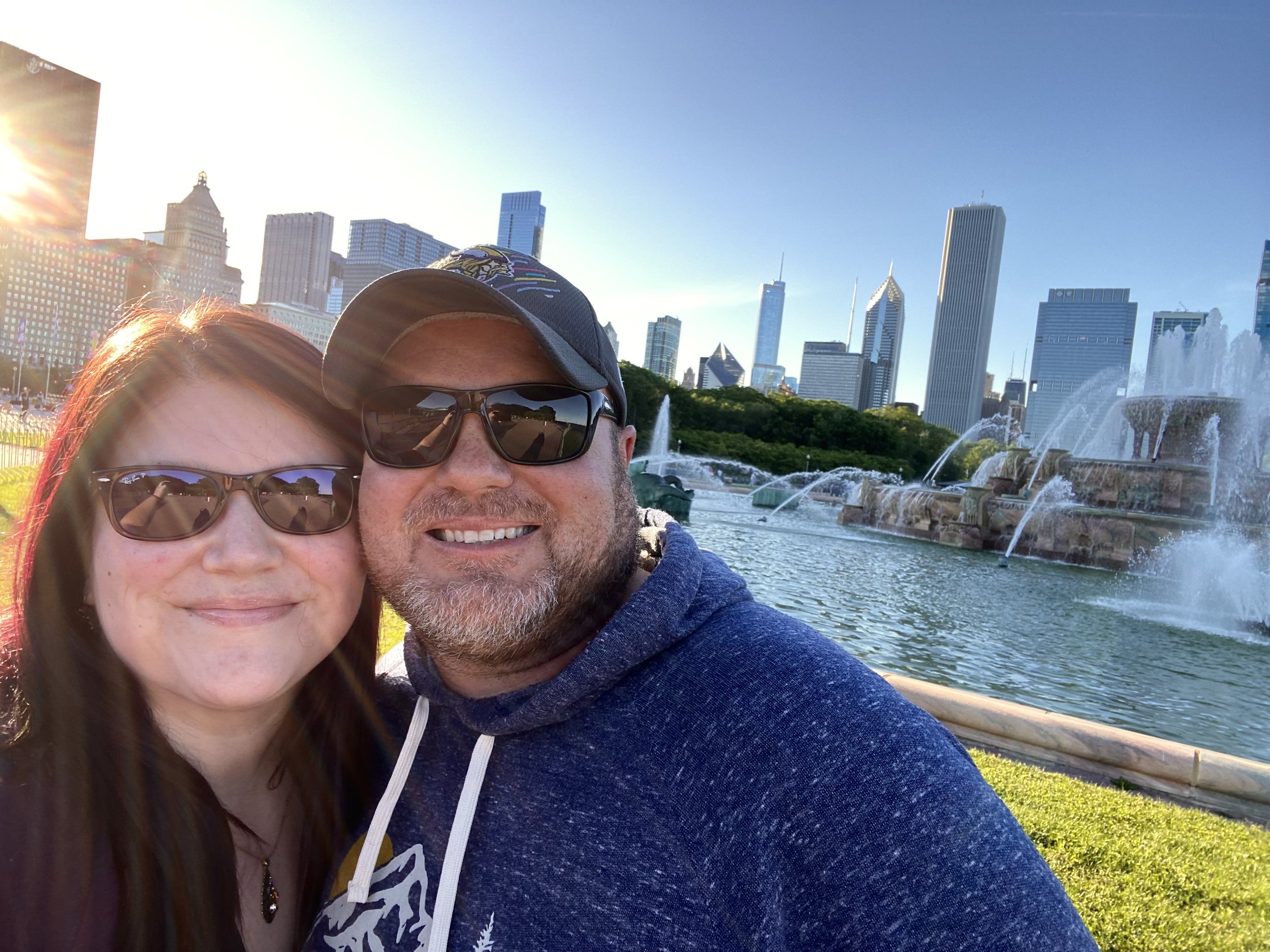 A smiling couple wearing sunglasses takes a selfie by the fountains at Millennium Park in Chicago with the city skyline in the background on a sunny day.