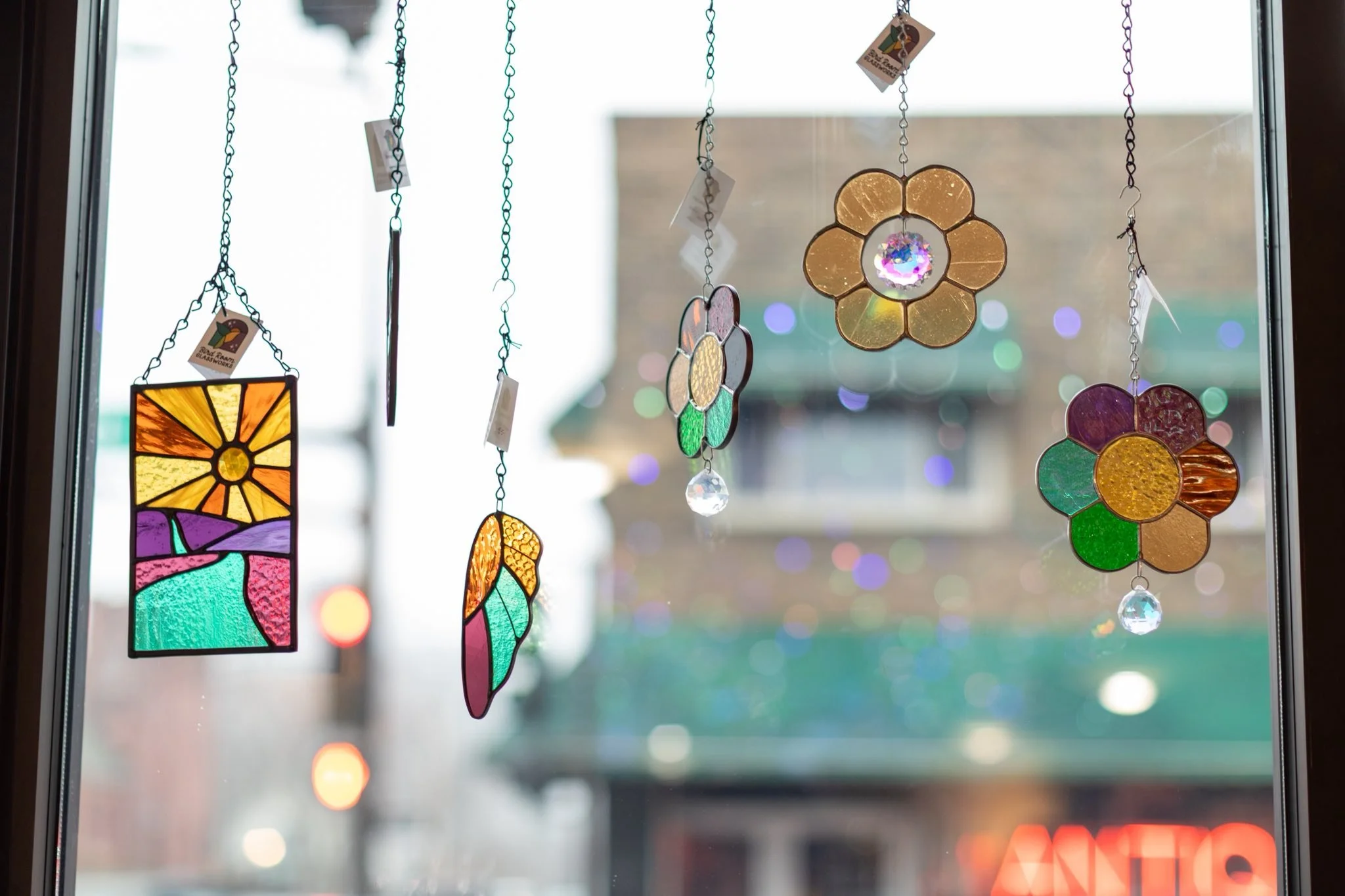 Colorful stained glass wind chimes hanging in front of a window with blurred city buildings and neon sign outside.