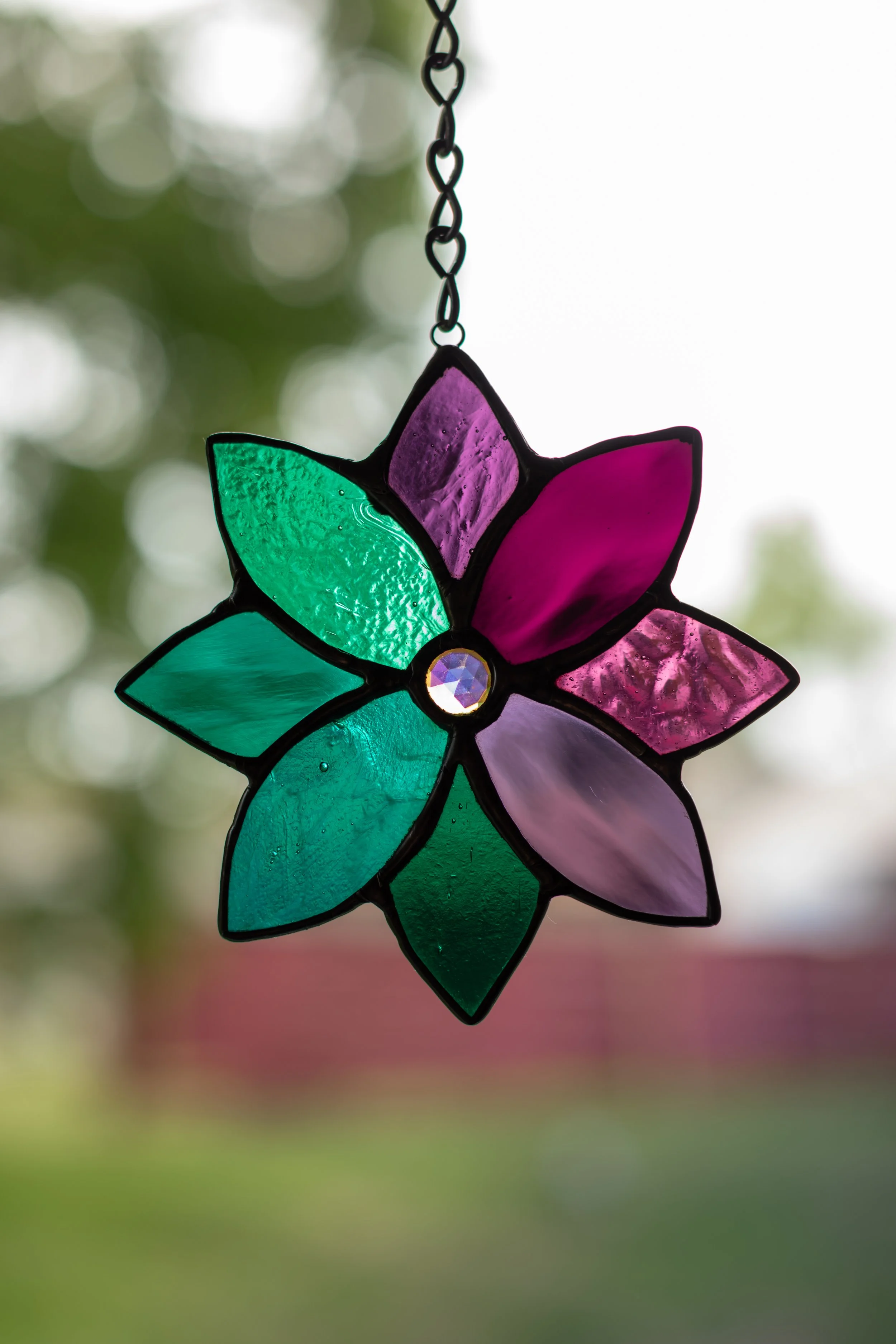 Colorful stained glass flower hanging from a chain, with green, purple, pink, and blue petals, against a blurred outdoor background.