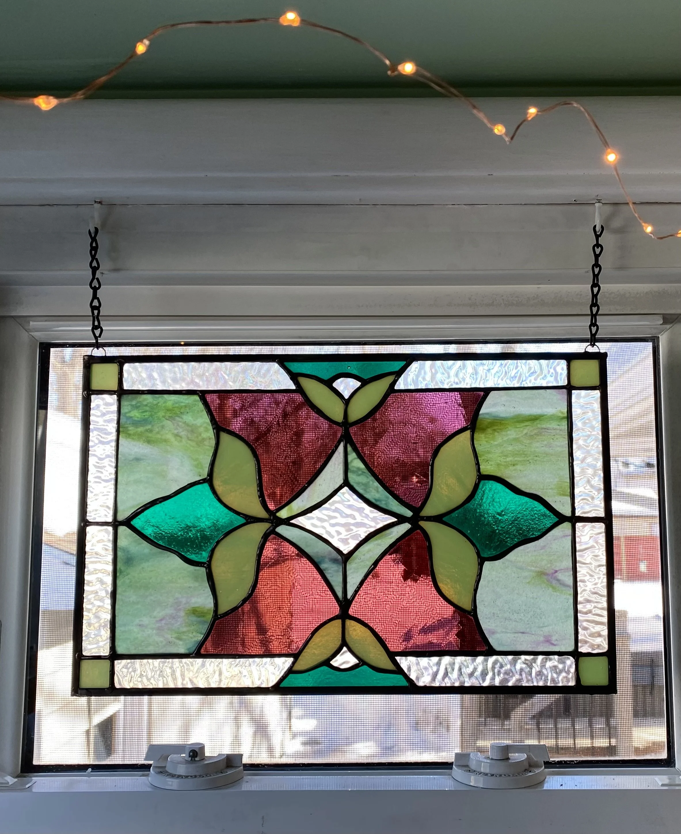 Colorful stained glass window with pink, green, and clear glass pieces in a geometric pattern, hanging in a window with a string of small orange lights above.
