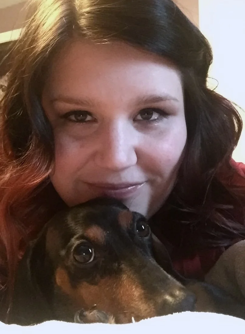 A woman with wavy dark hair and a slight smile taking a selfie with her dog, a black and tan dachshund, close to her face.