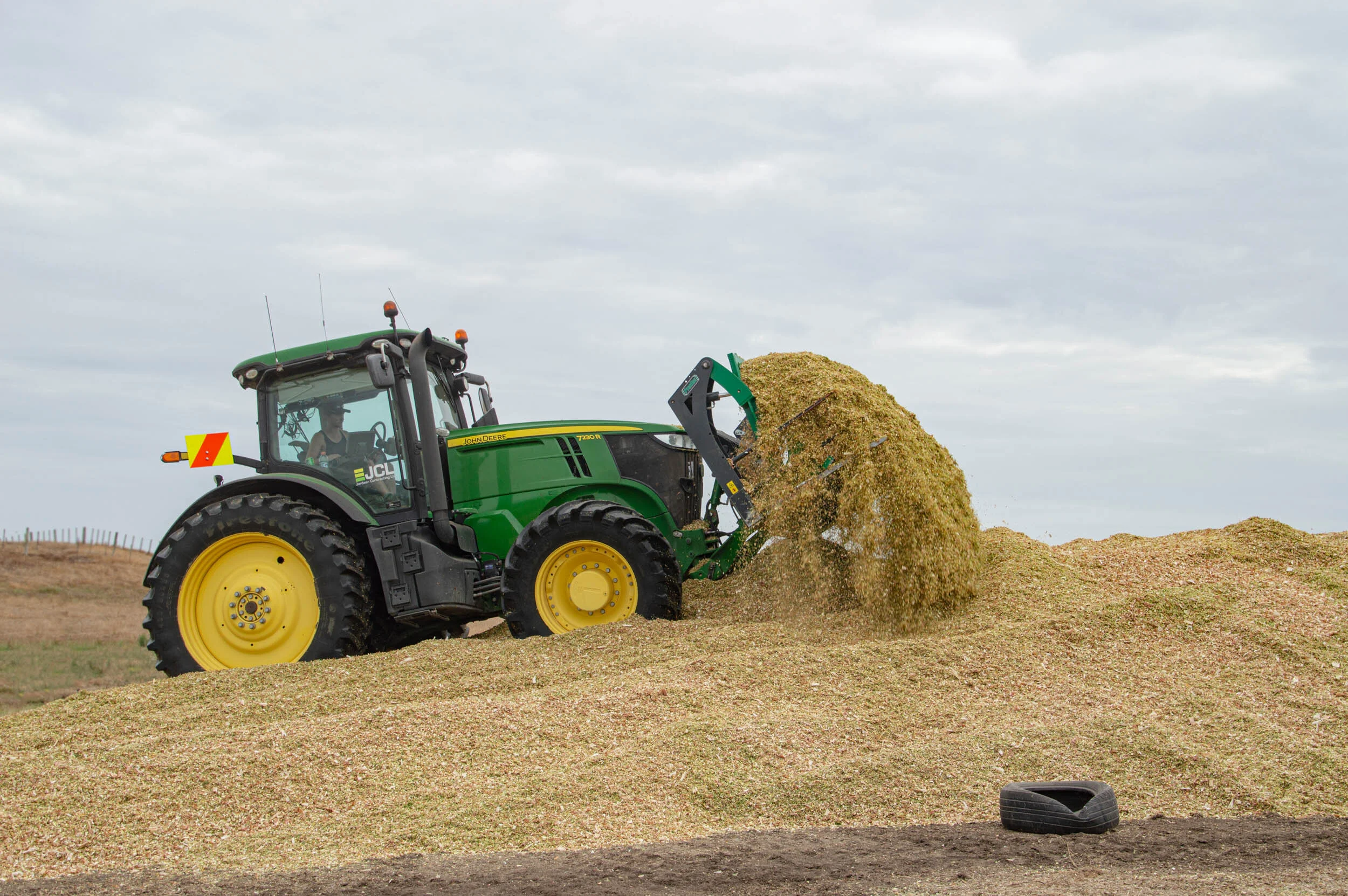 Green John Deere tractor with yellow wheels spreading a pile of grain or silage on a farm field.