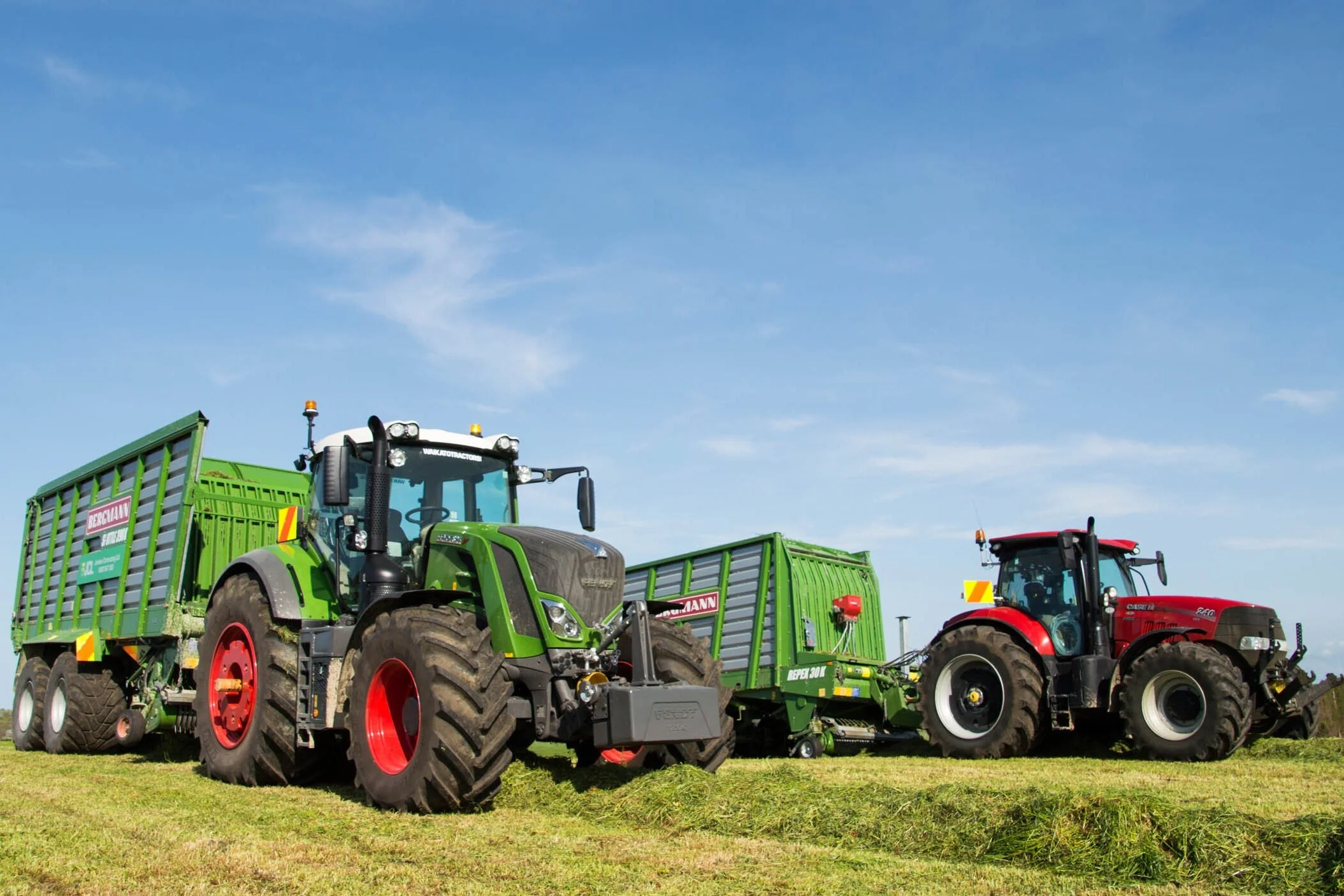 Two large farm tractors, one green and one red, are working in a field under a blue sky, with green grass visible in the foreground.