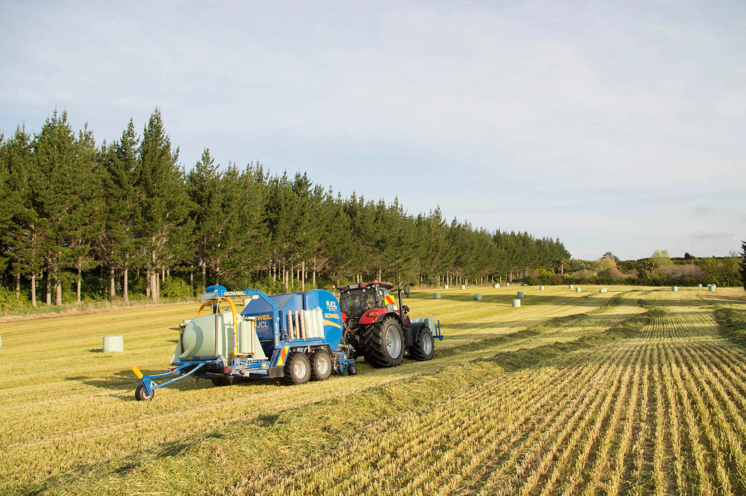 A tractor pulling a large piece of equipment working on a field of crops under a partly cloudy sky, with a row of trees in the background and hay bales scattered across the field.