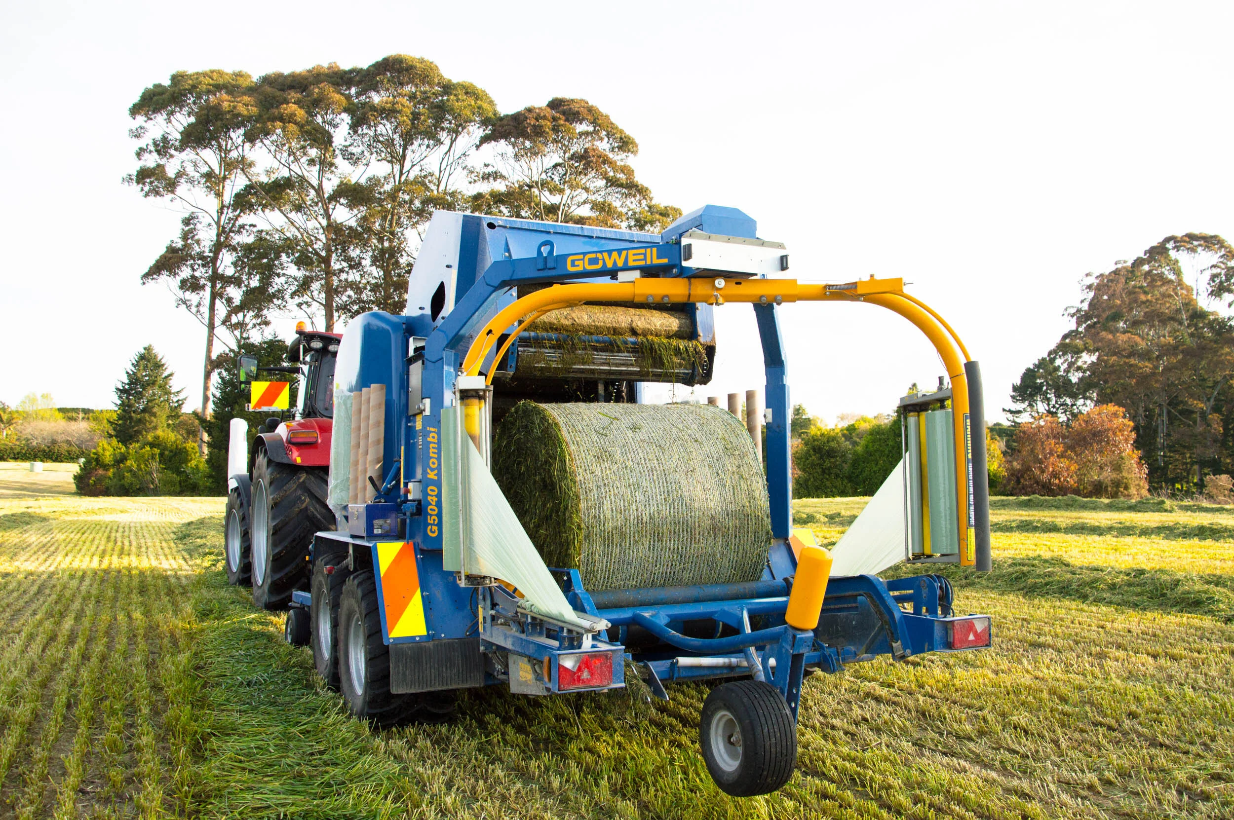 A tractor with a bale wrapper attachment wrapping a large round hay bale in a field during daytime.